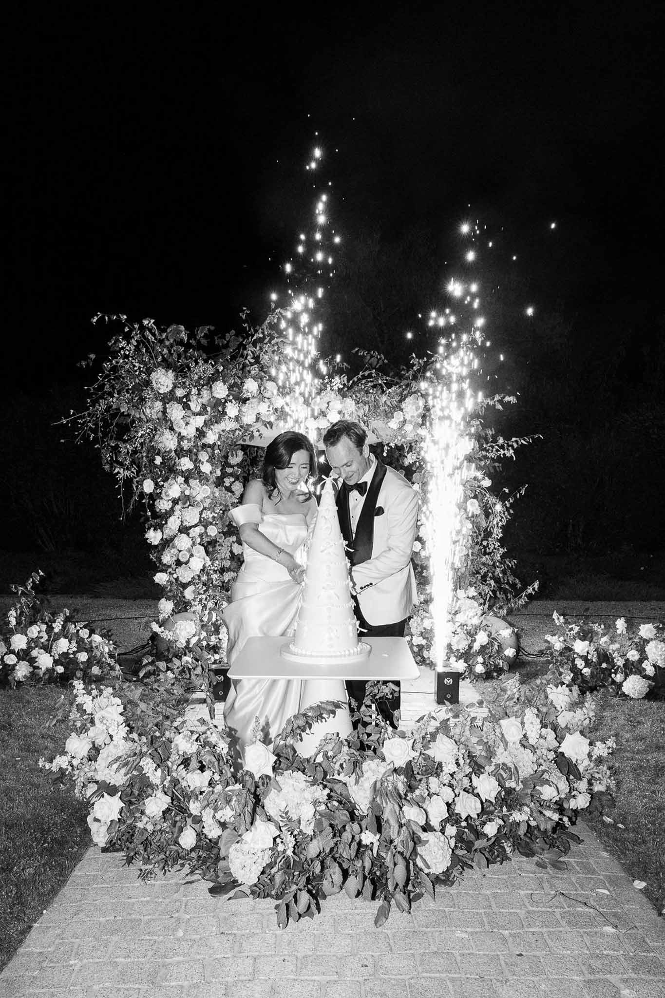 Black and white shot of couple cutting multi-tiered cake with sparkler fountains and lush floral arch behind them