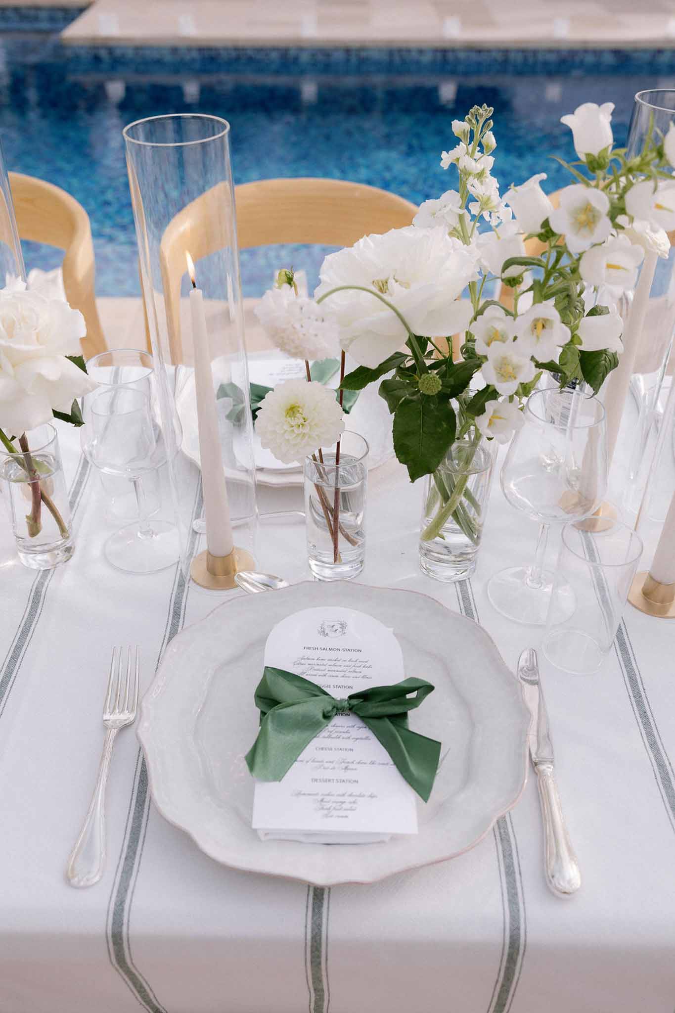 Place setting with scallop-edged plate, green ribbon menu card, white dahlia bud vases, and brass candle holders