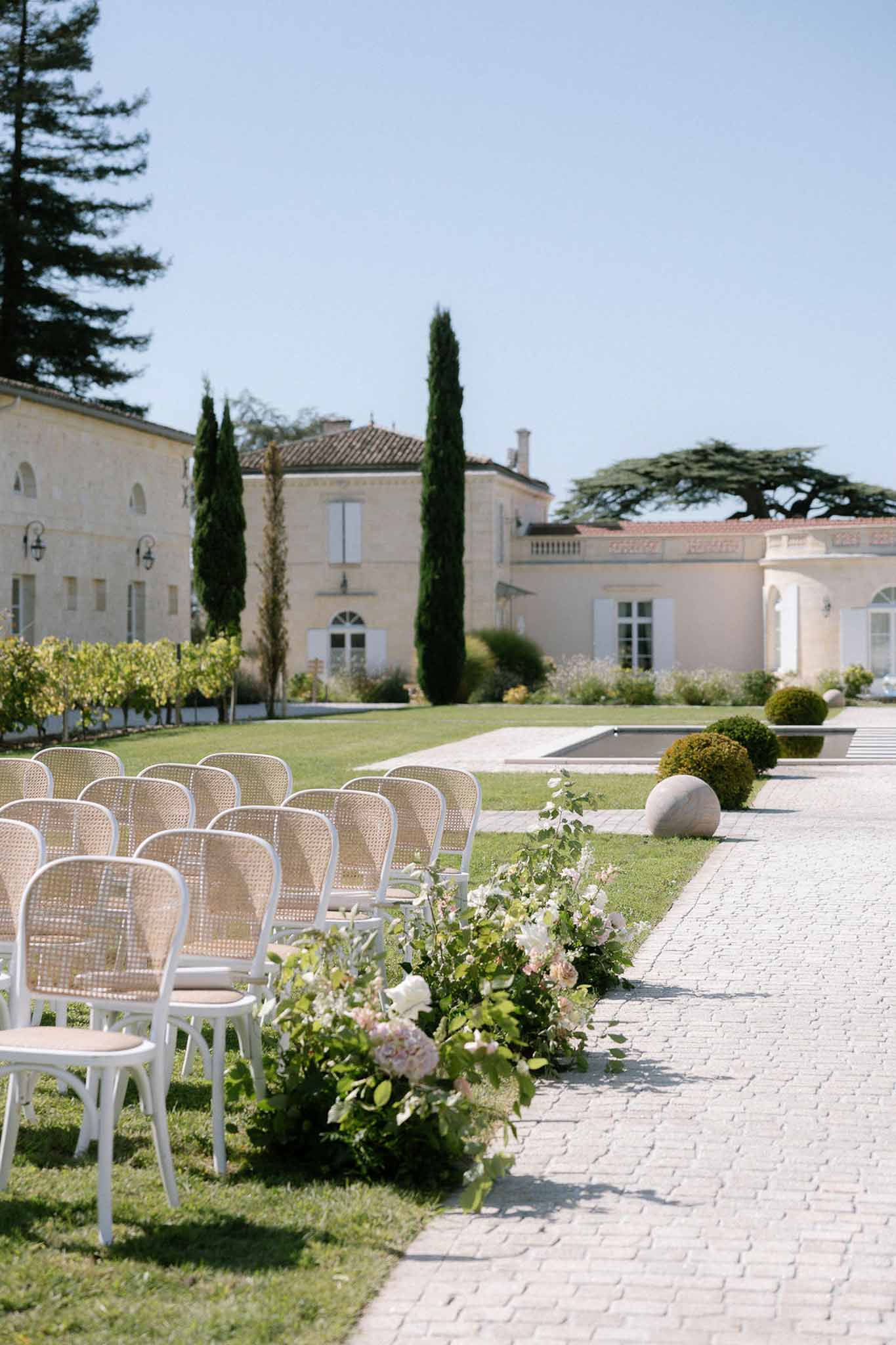 Outdoor ceremony setup at French chateau with white cane-back chairs, blush hydrangea aisle flowers and reflecting pool