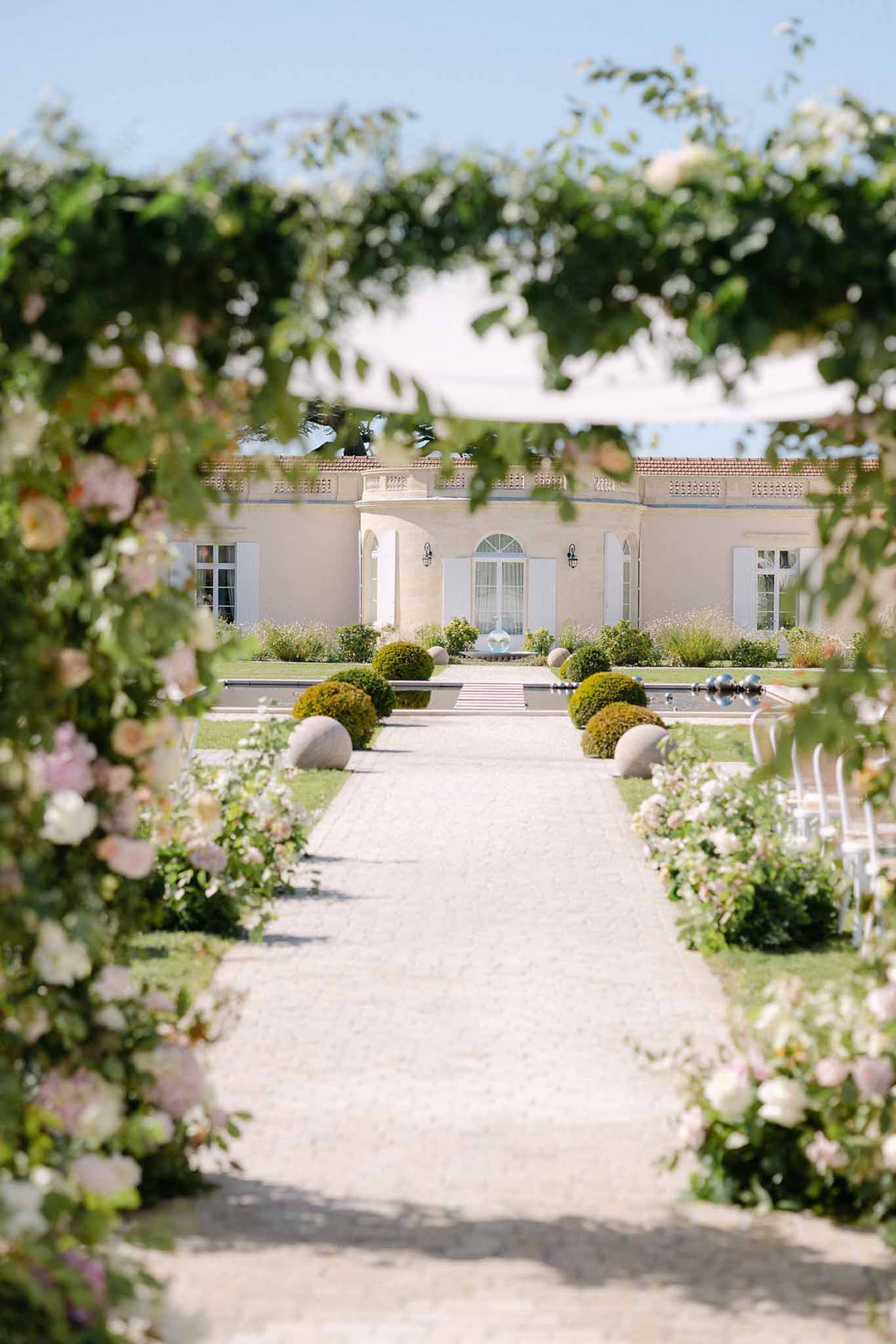 Ceremony aisle framed through blush rose arch toward chateau facade with boxwood and reflecting pool