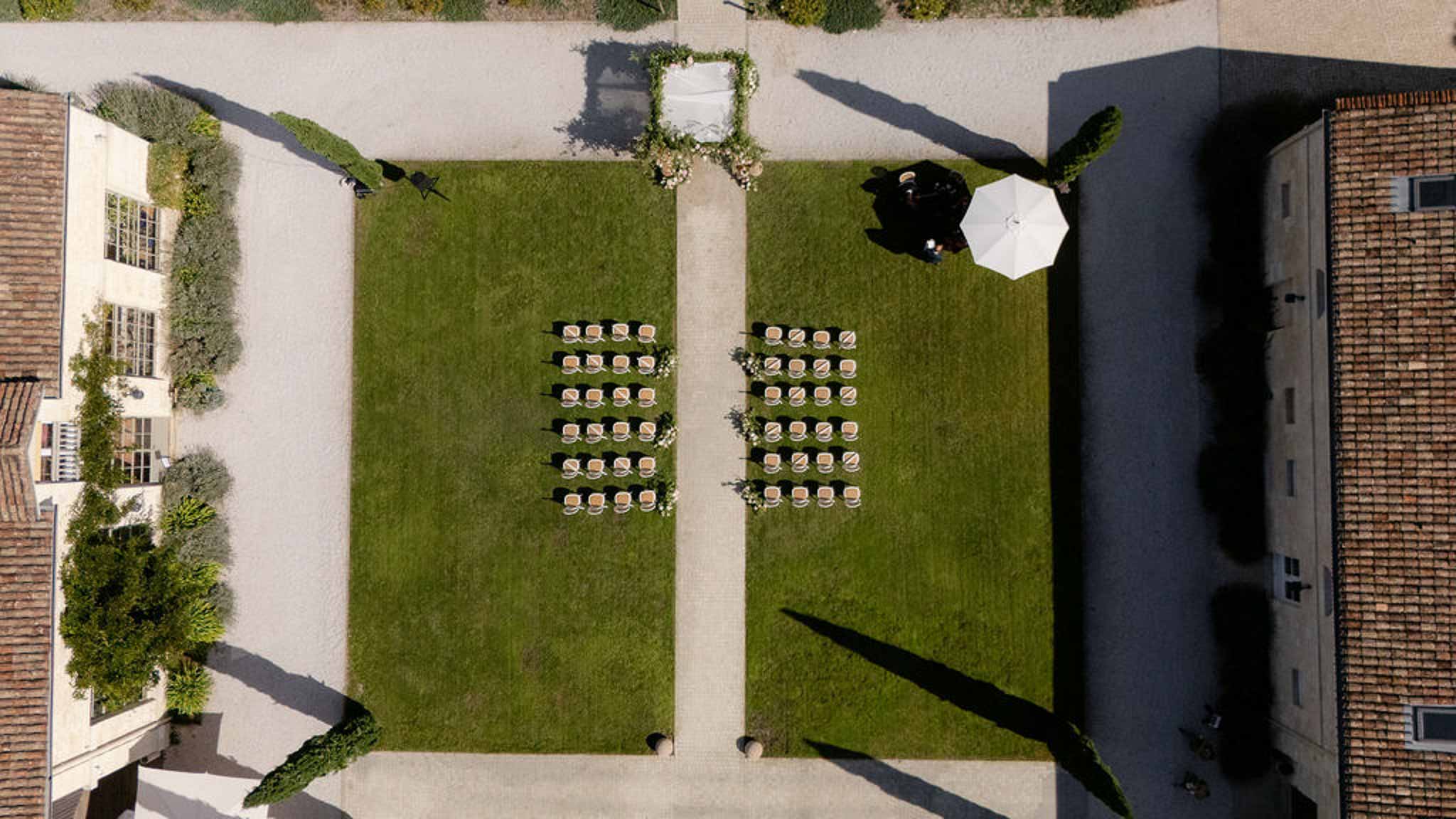 Aerial of white chair ceremony setup on lawn between terracotta-roofed buildings with floral arch