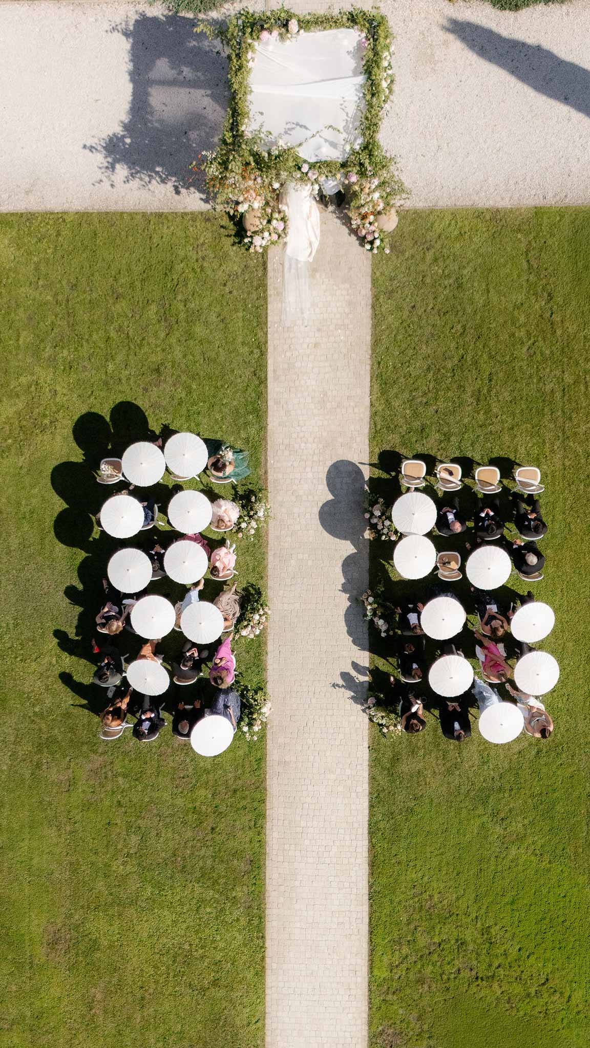 Aerial drone view of outdoor wedding ceremony on lawn with white parasols over guest seating and greenery-covered arch