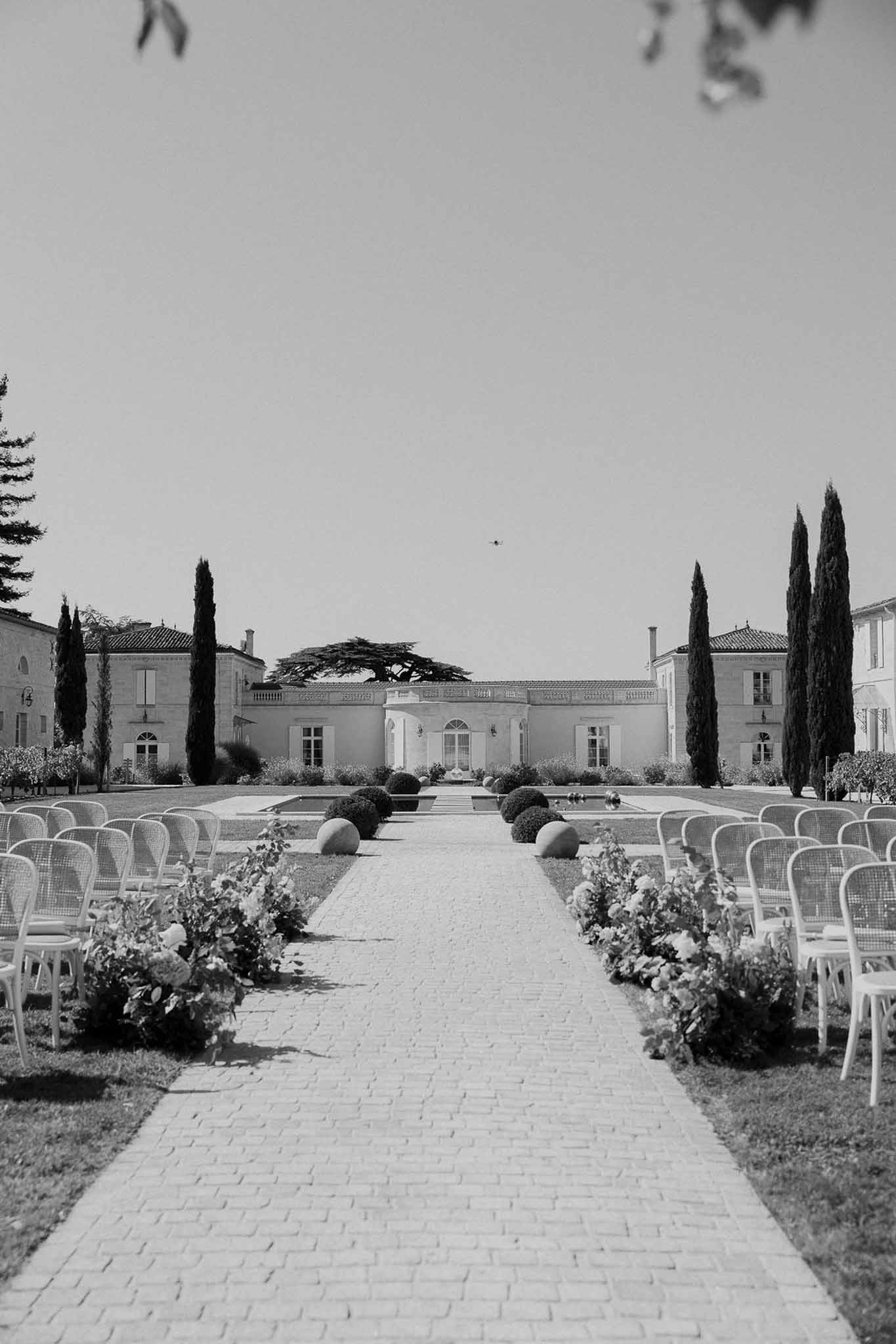 Black and white ceremony setup with cane chairs, floral aisle clusters, and reflecting pool before chateau