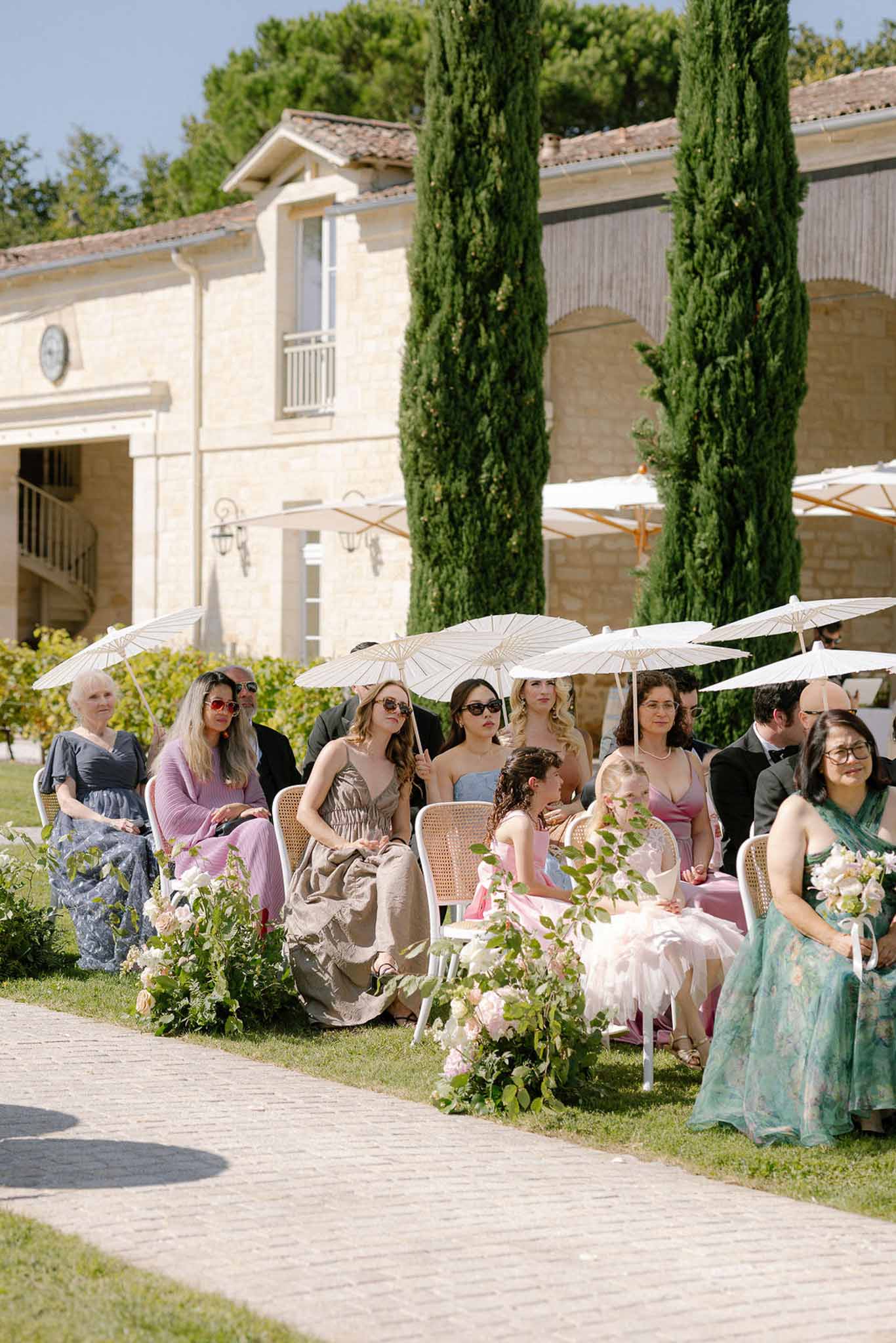 Guests with white parasols in rattan chairs on lawn with blush rose aisle arrangements before honey-stone building