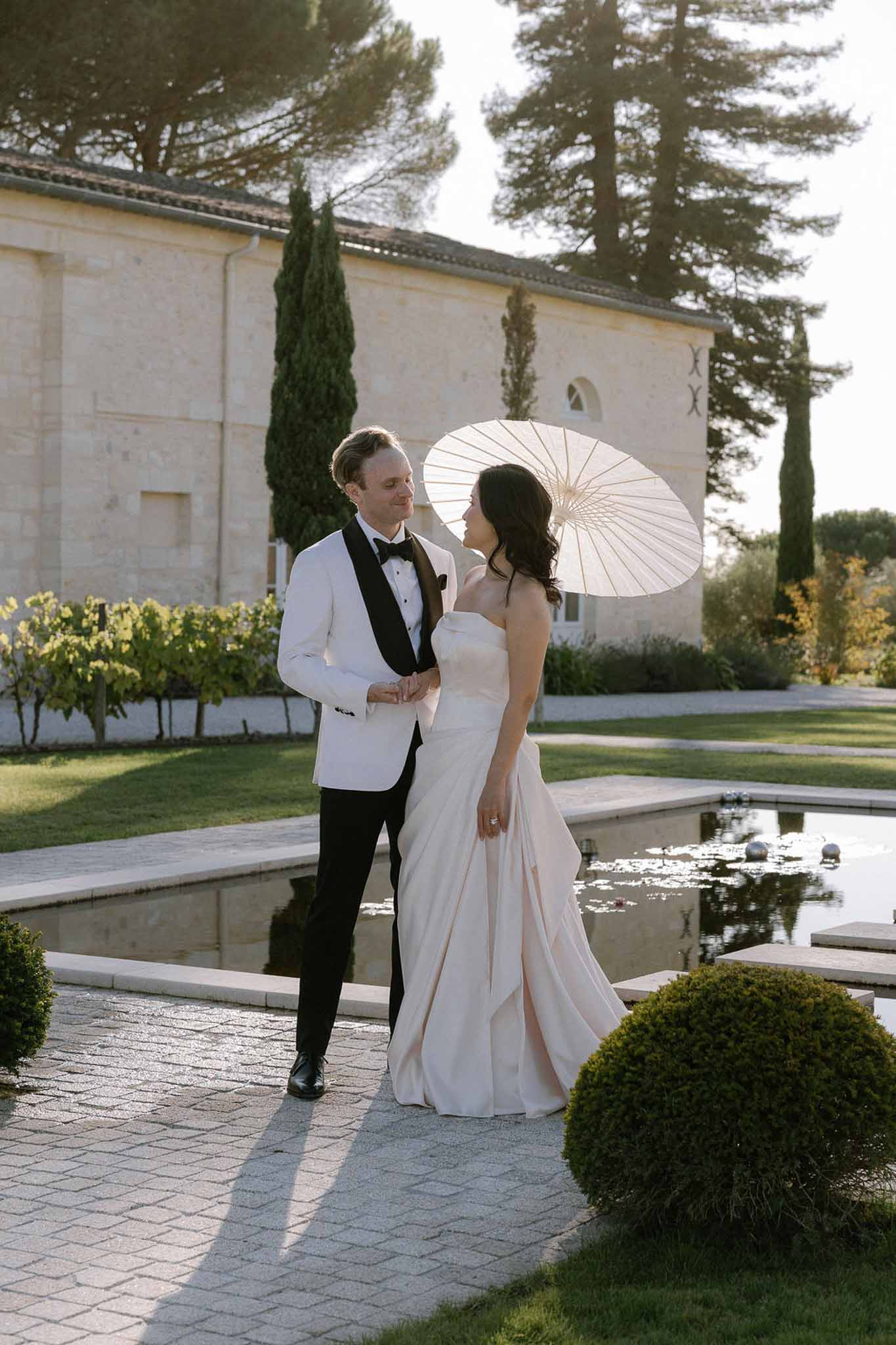 Bride in blush-pink gown with parasol and groom in white dinner jacket walking beside reflecting pool at stone estate