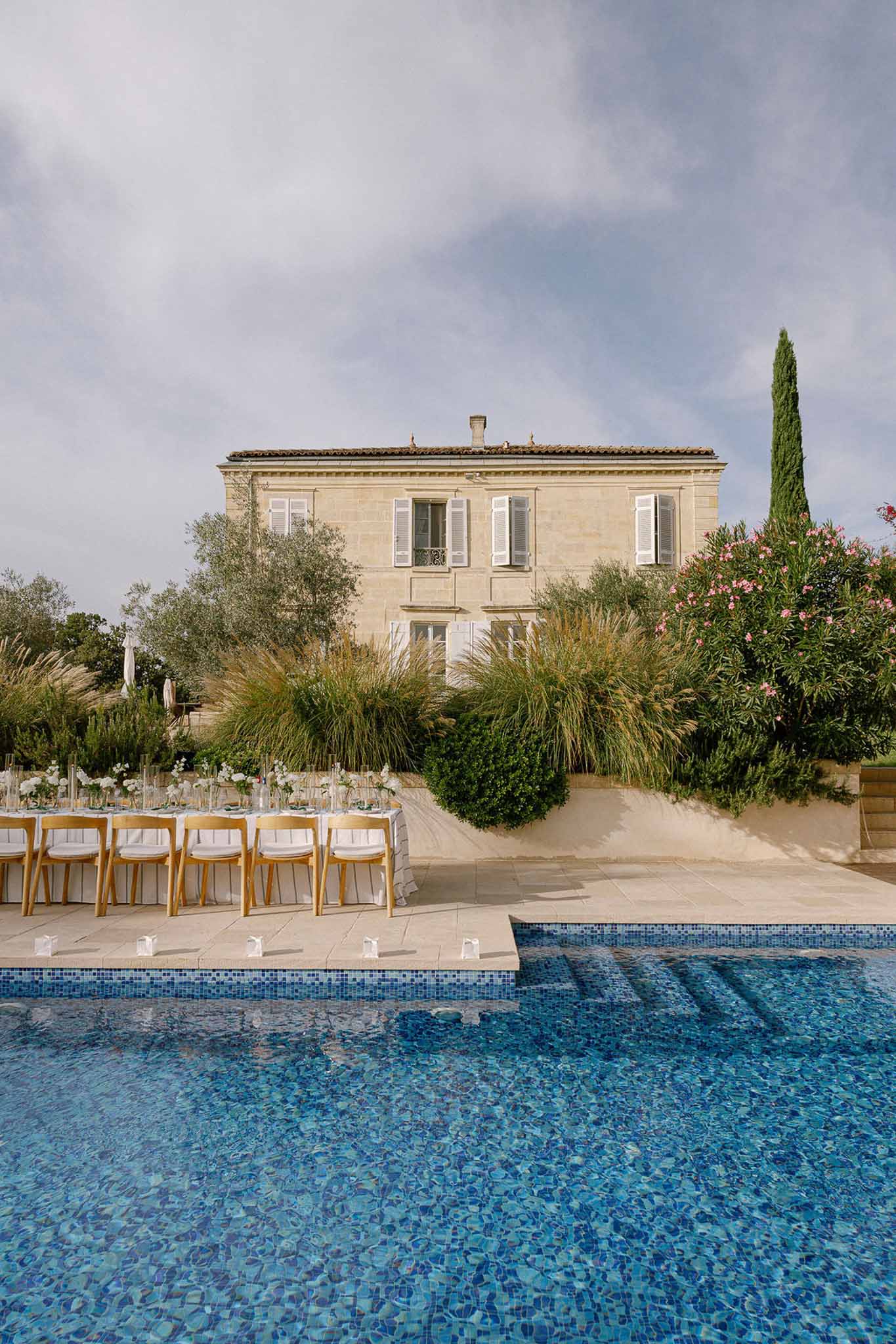 Long reception table with white linens and floral centerpieces set poolside at limestone manor house