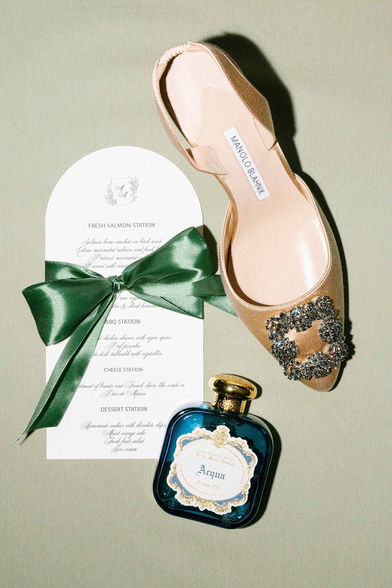 Manolo Blahnik champagne slingback with crystal buckle beside arch menu card and teal perfume on sage surface