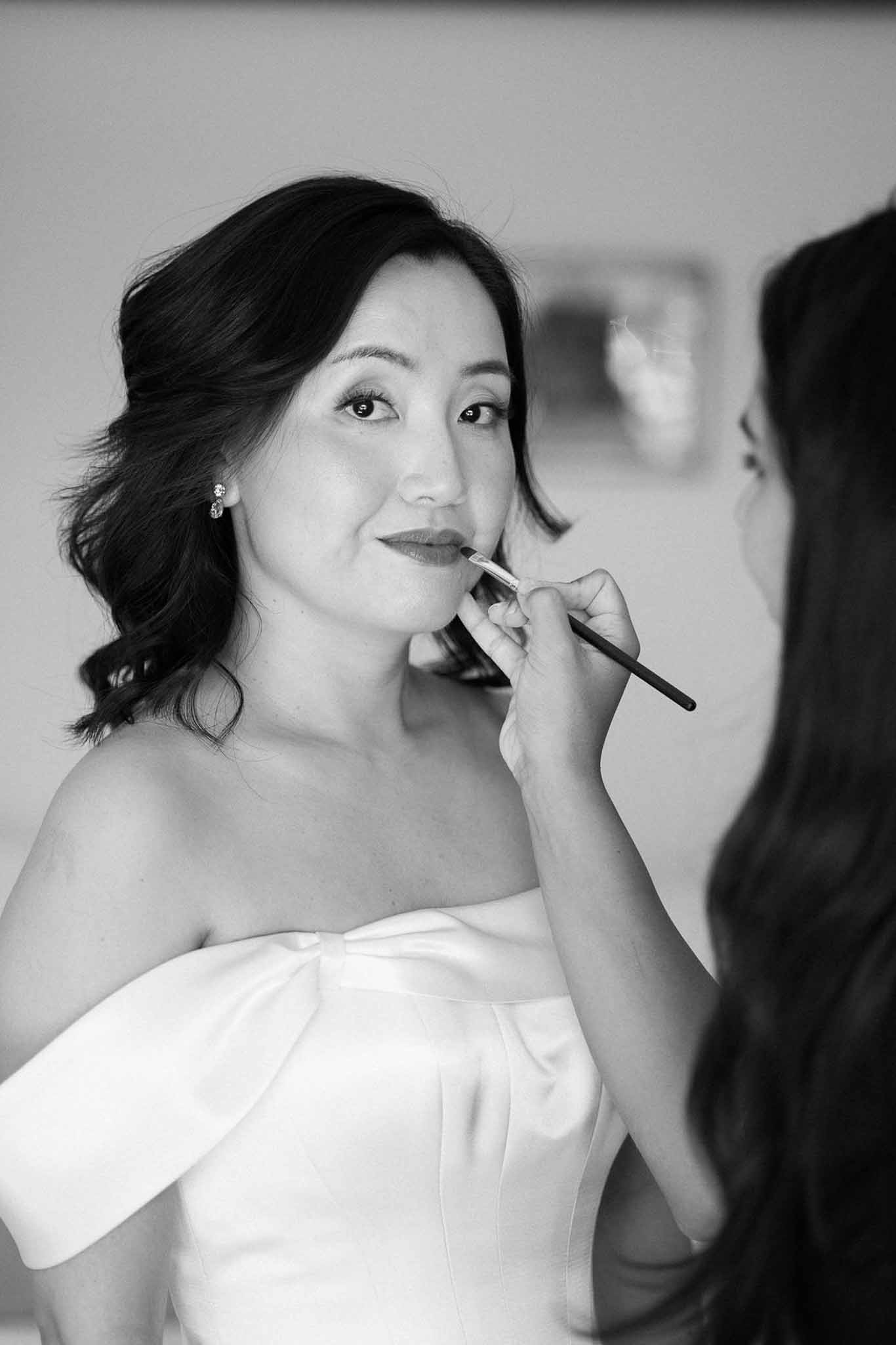Black and white close-up of makeup artist applying lipstick to bride in strapless off-shoulder gown