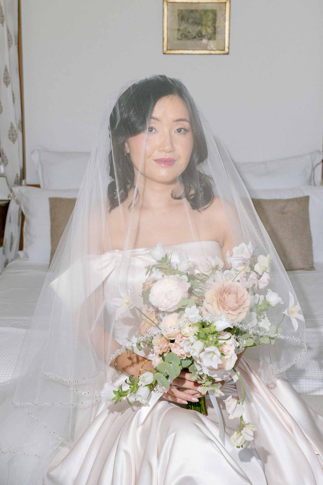 Bride seated on bed in satin ballgown with beaded-edge veil holding blush and peach rose bouquet hazy film quality