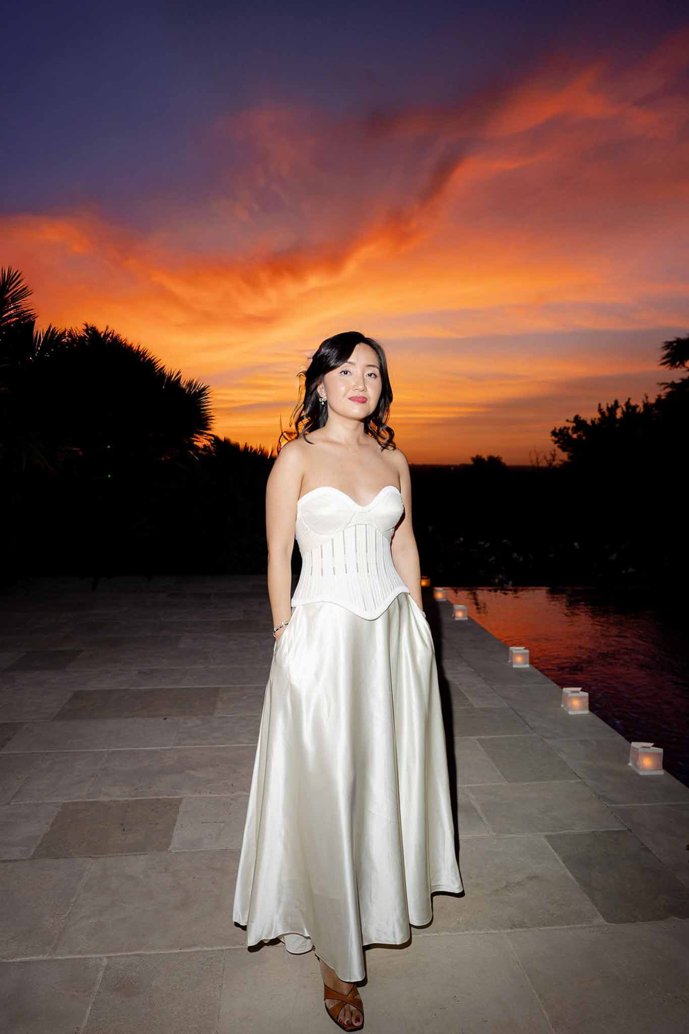 Bride in two-piece ivory corset and satin skirt on poolside terrace with candle lanterns at dusk