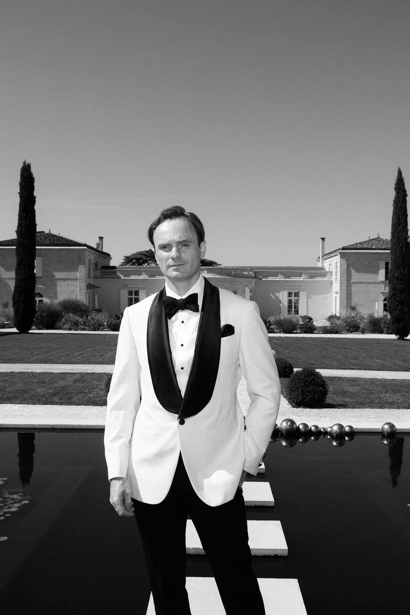 Black and white portrait of groom in white dinner jacket standing before chateau reflecting pool and topiary