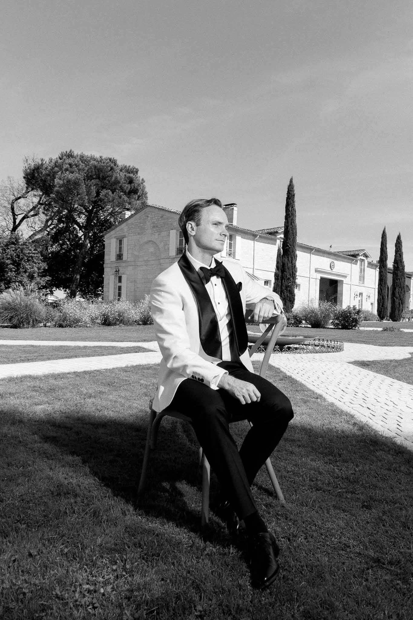 Black and white groom in white dinner jacket seated on lawn with chateau facade cypress trees and garden borders behind