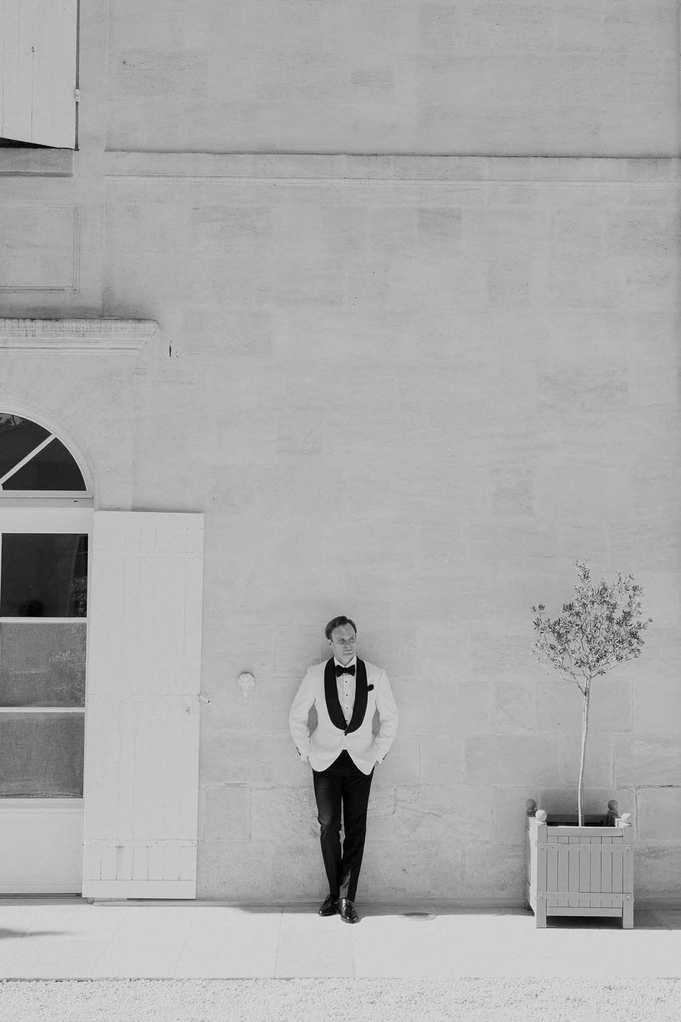 Black and white full-length portrait of groom in white dinner jacket standing against chateau stone facade