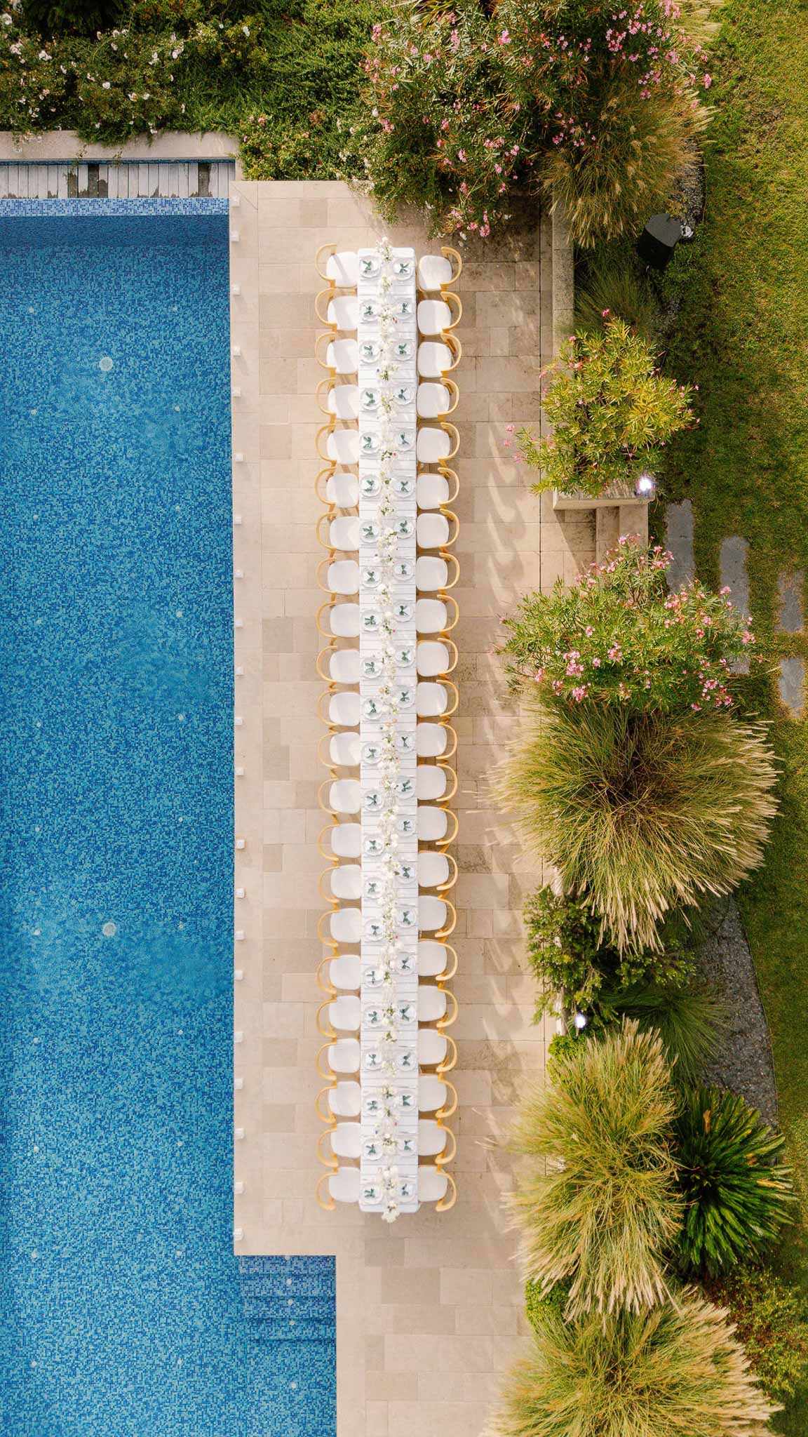 Aerial view of long white-linen banquet table with gold chairs set beside blue mosaic pool