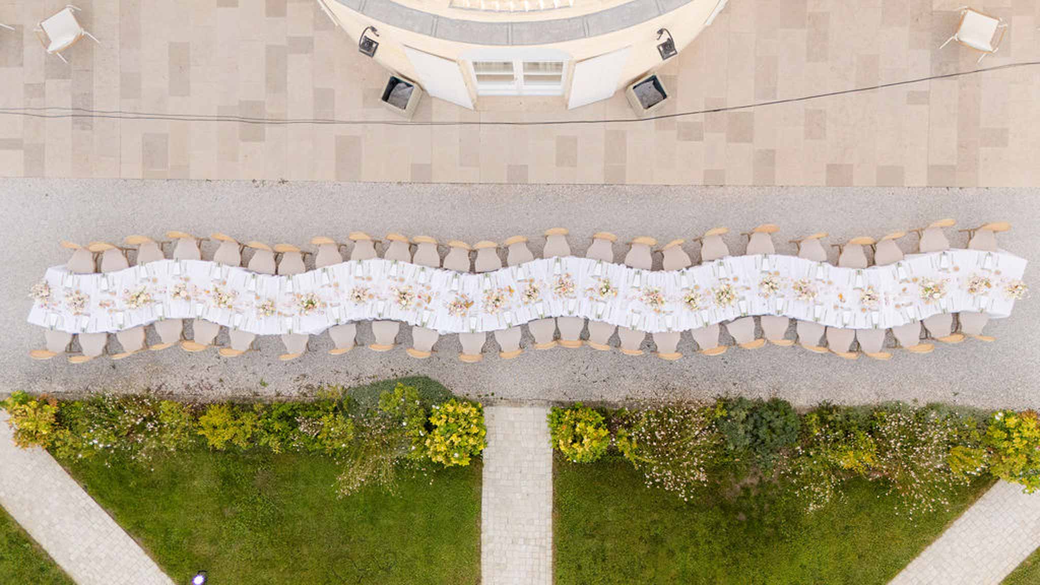 Aerial view of long serpentine feasting table with blush florals and cross-back chairs on courtyard