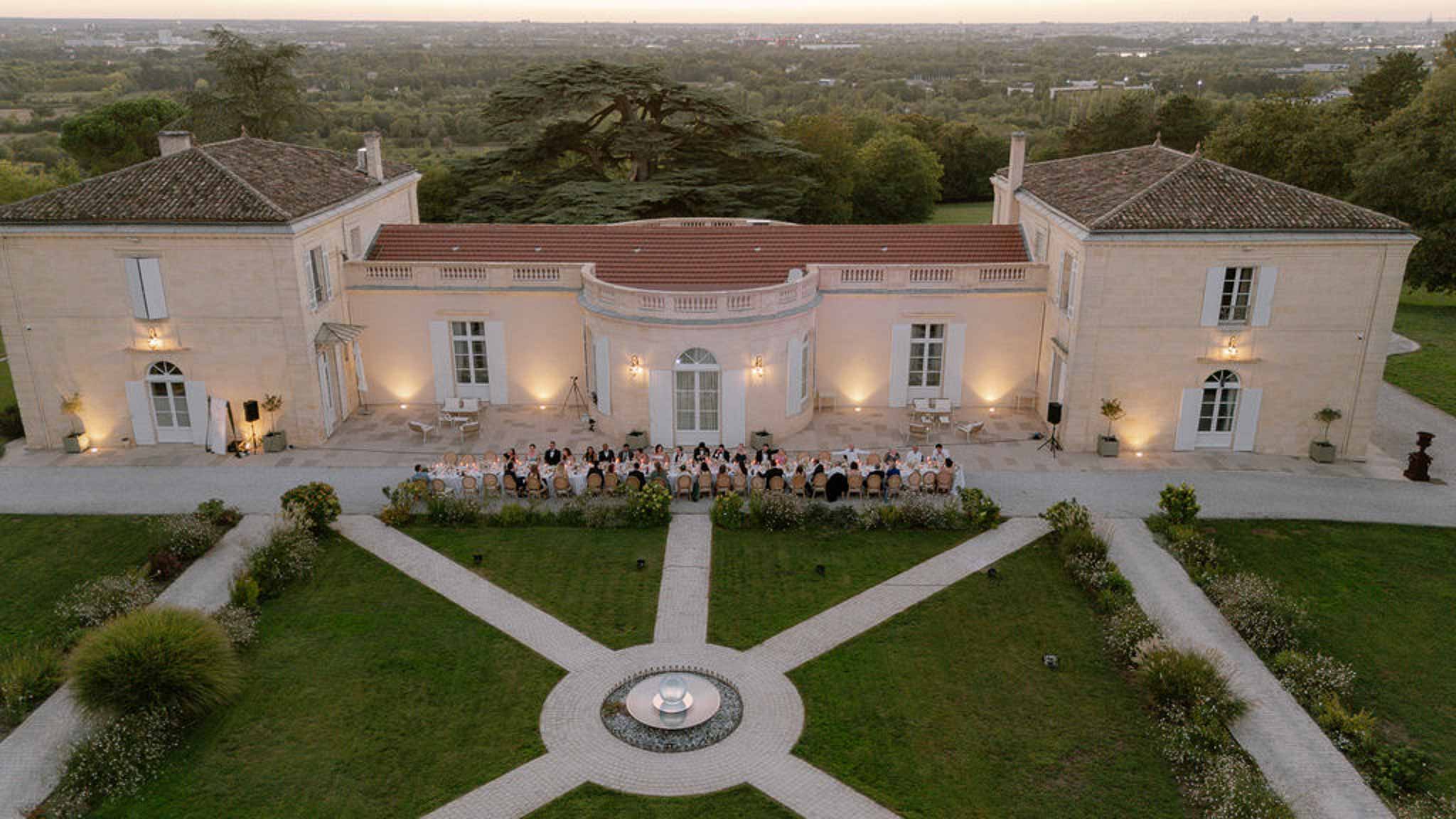 Aerial dusk view of 50 guests at long table before limestone chateau with geometric garden and fountain