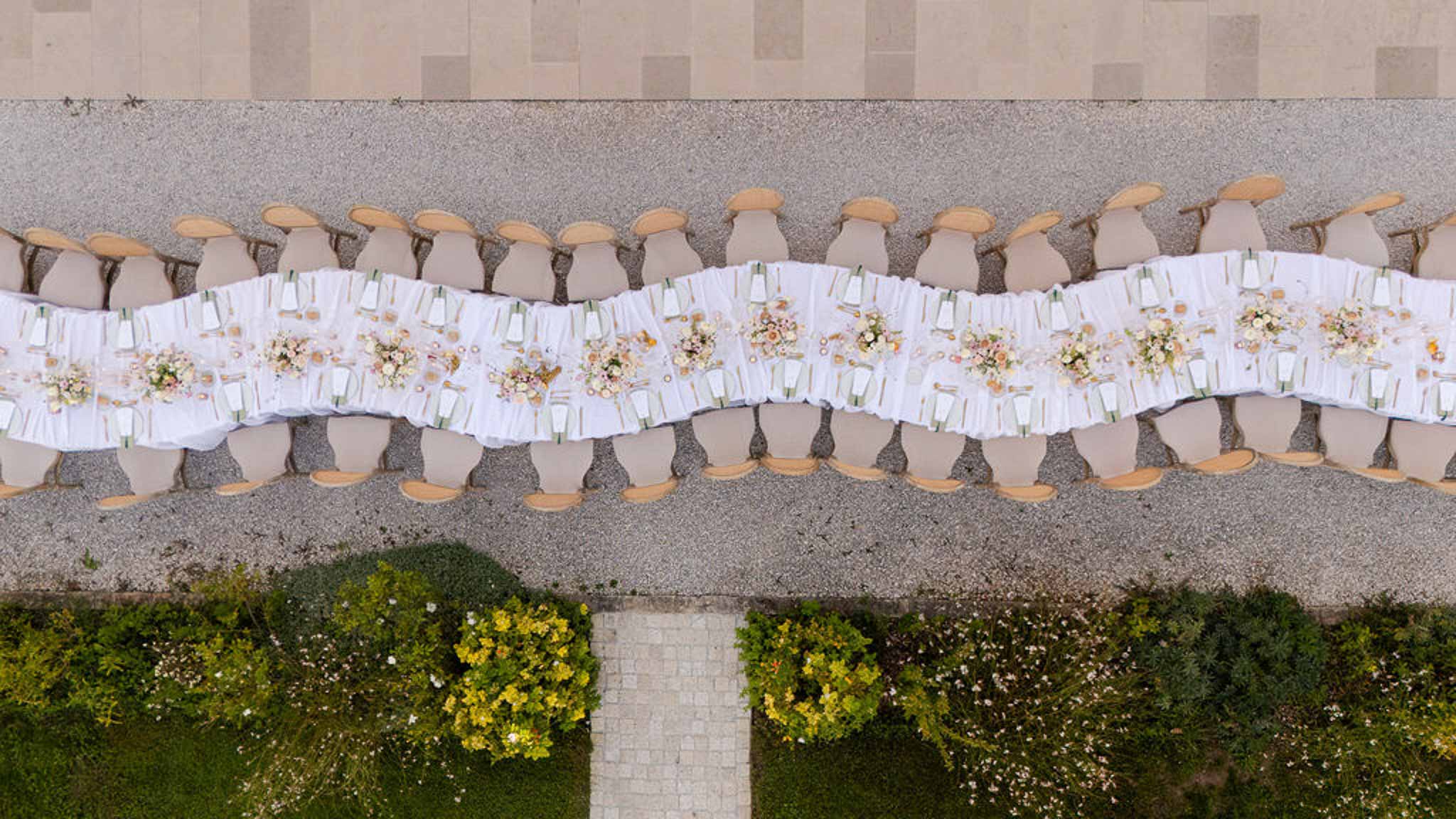 Aerial view of long reception table with blush rose centrepieces and sage menu cards on gravel courtyard