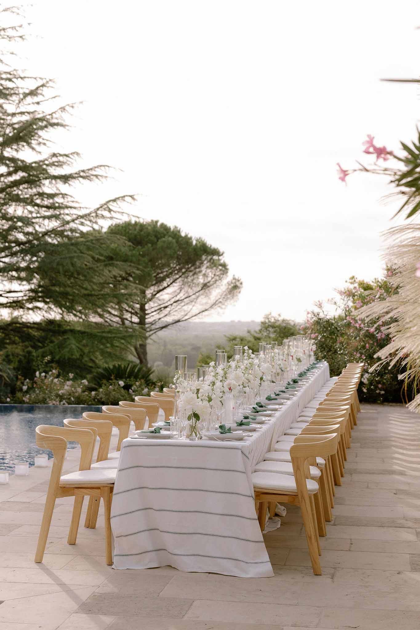 Long reception table beside swimming pool with white floral centerpieces, glass cylinder vases, and wood chairs