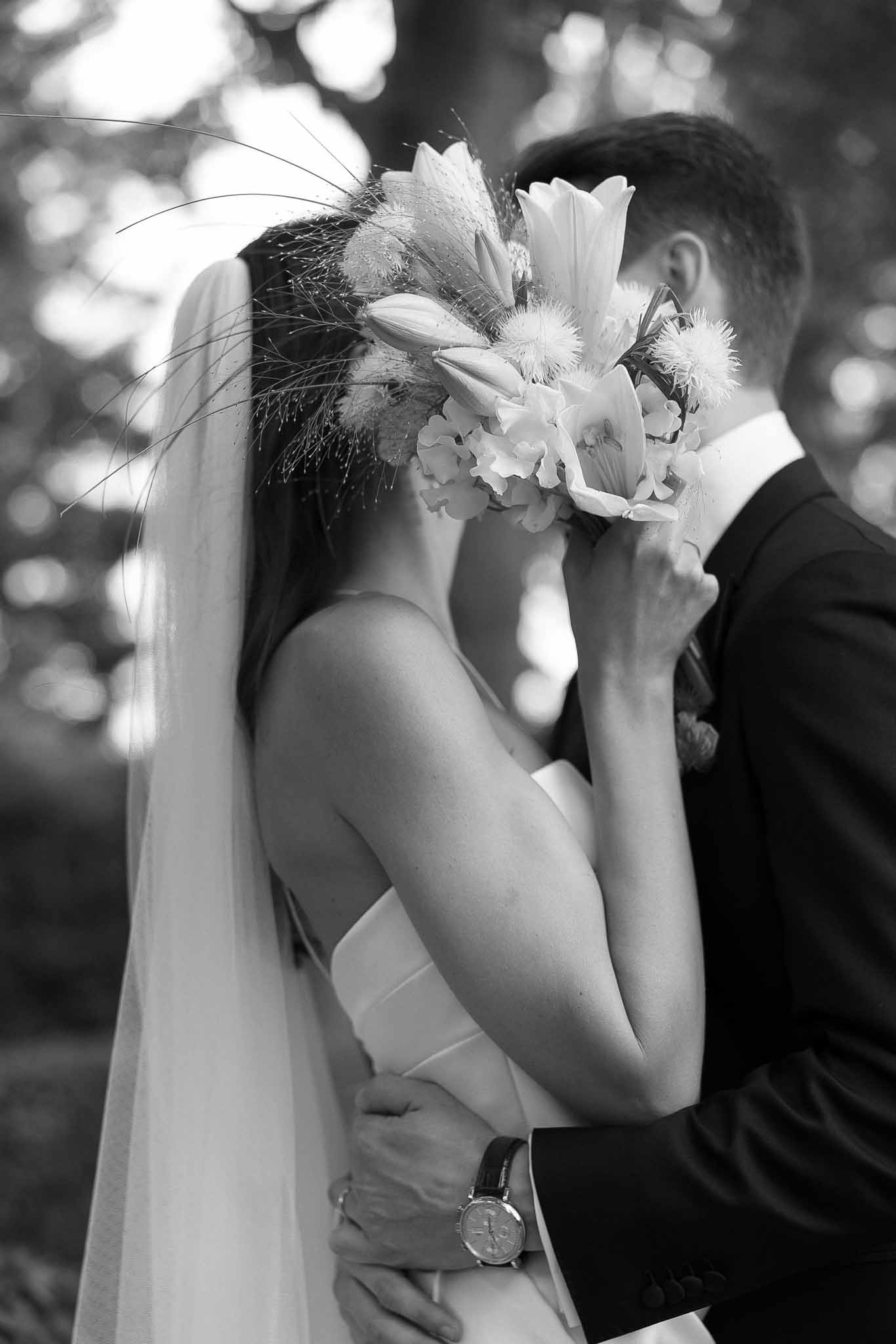 Black and white close-up of couple embracing behind organic bridal bouquet with lilies and dried grasses