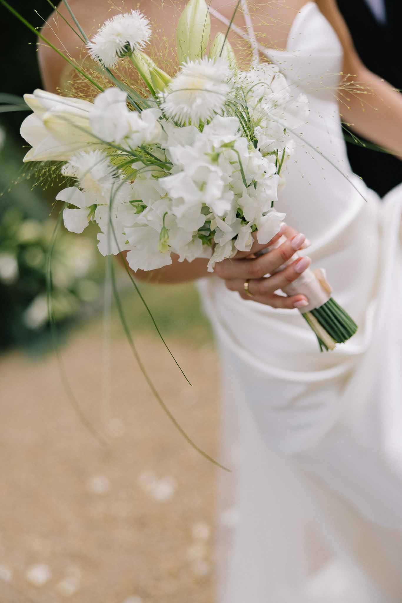 Close-up of bridal bouquet with white sweet peas, lilies, and trailing golden grasses against satin gown