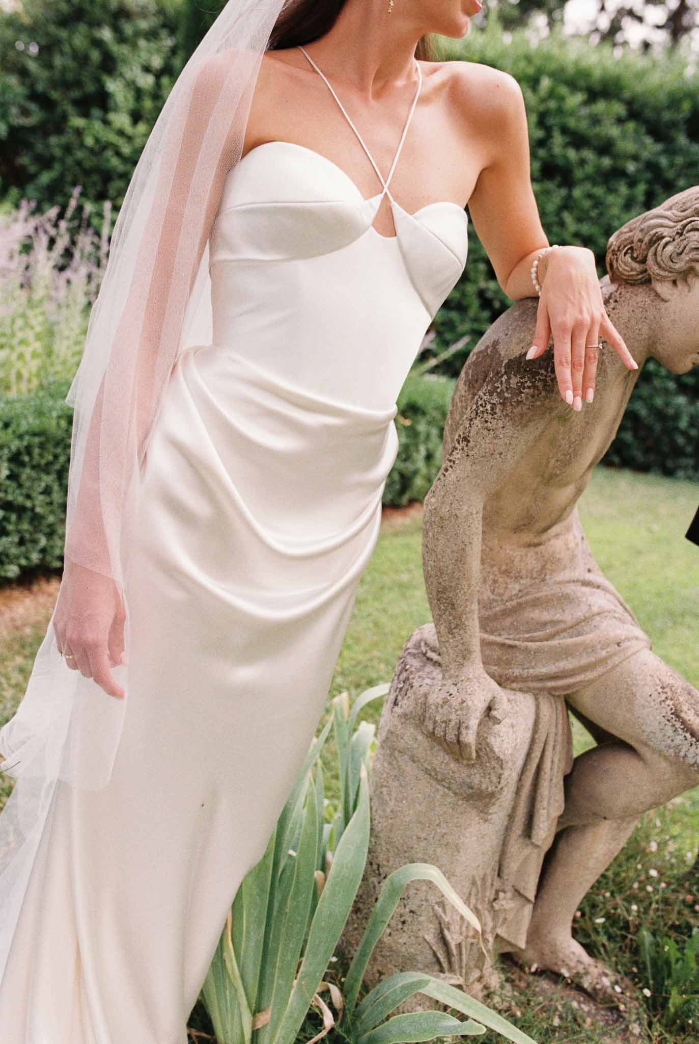 Bride in fitted ivory slip gown with sheer veil leaning on weathered classical garden sculpture