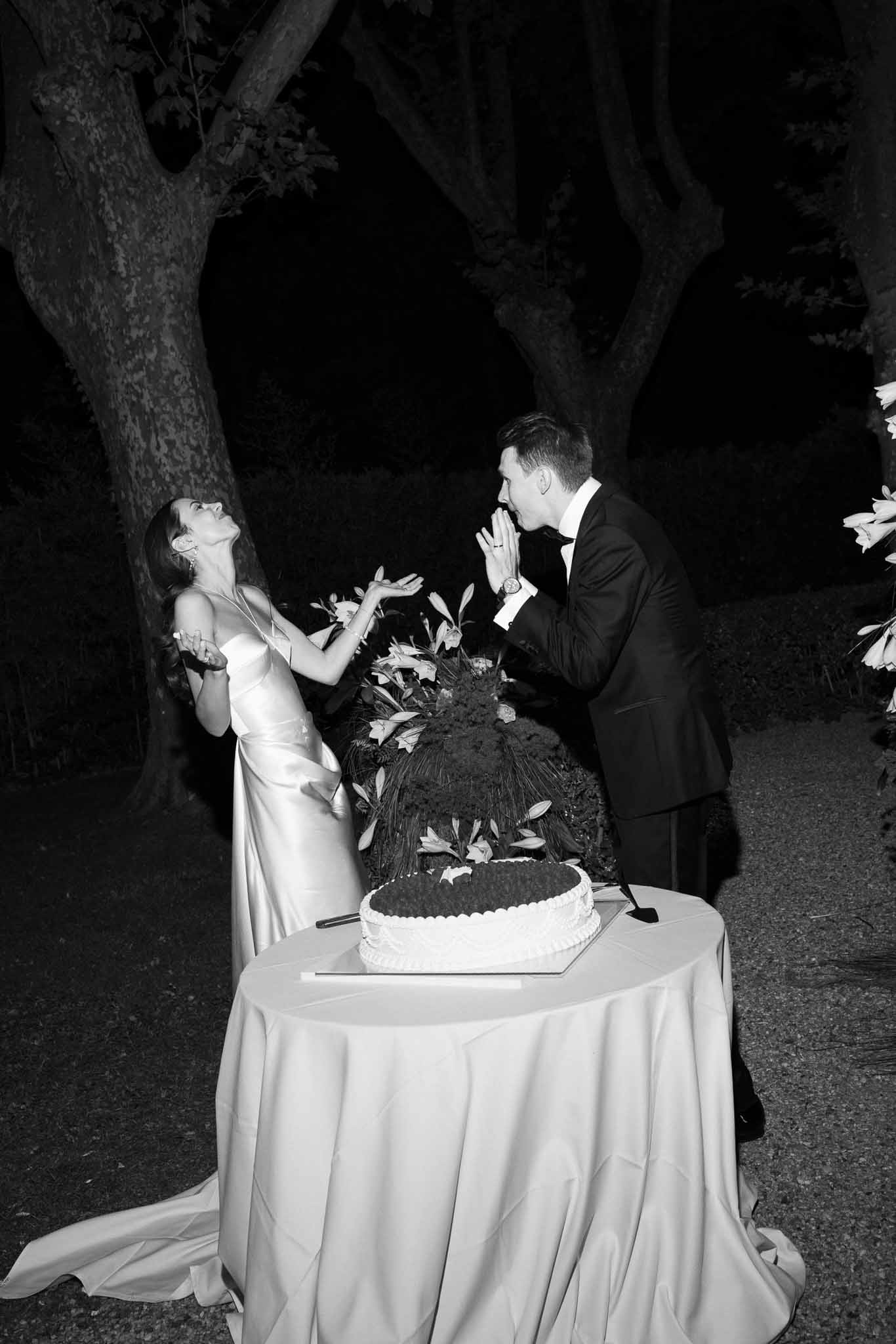 Black and white cake cutting with laughing bride and amused groom beside two-tier lily-topped cake at night
