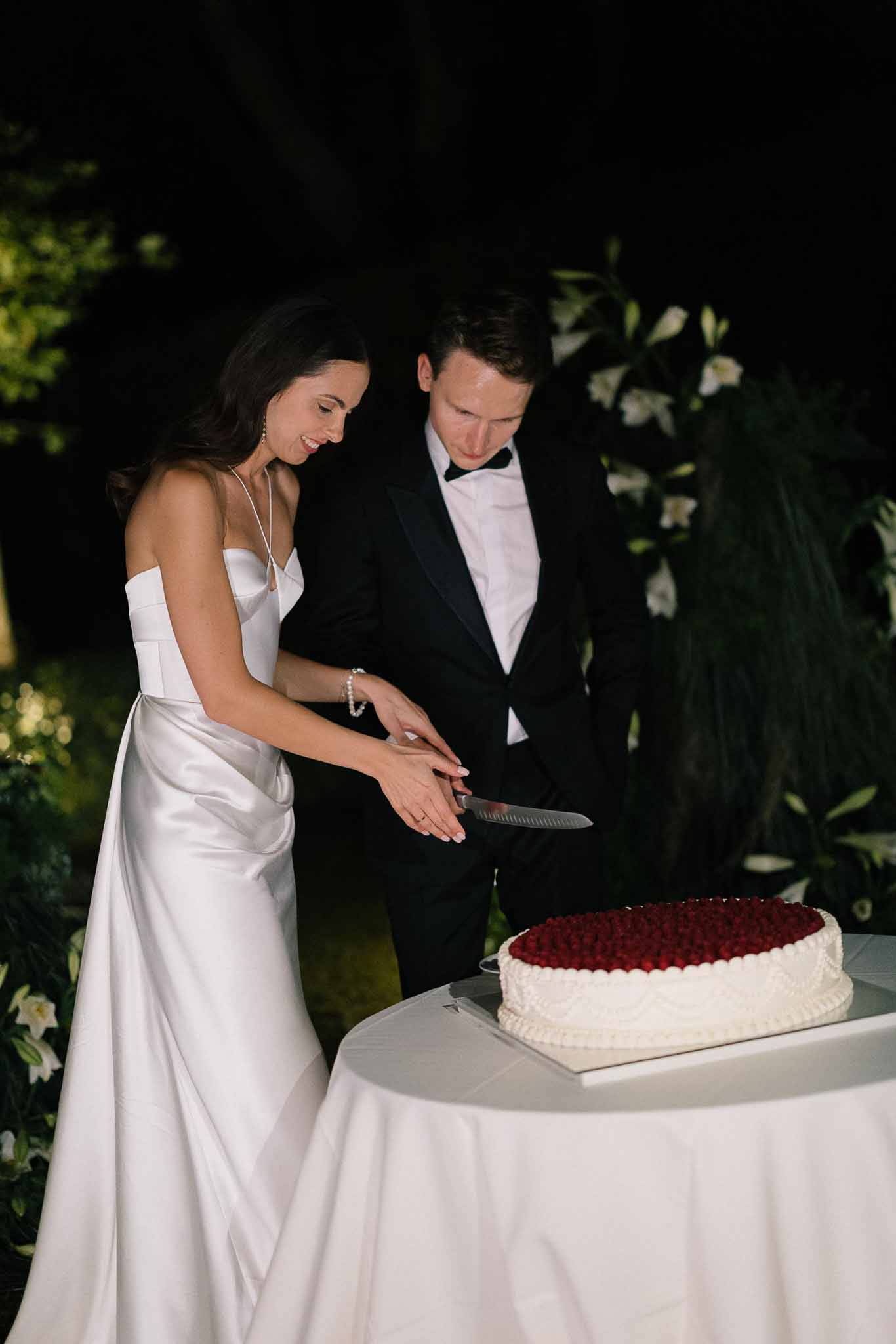 Couple cutting raspberry-topped single-tier cake bride in ivory satin slip gown groom in black tuxedo at night