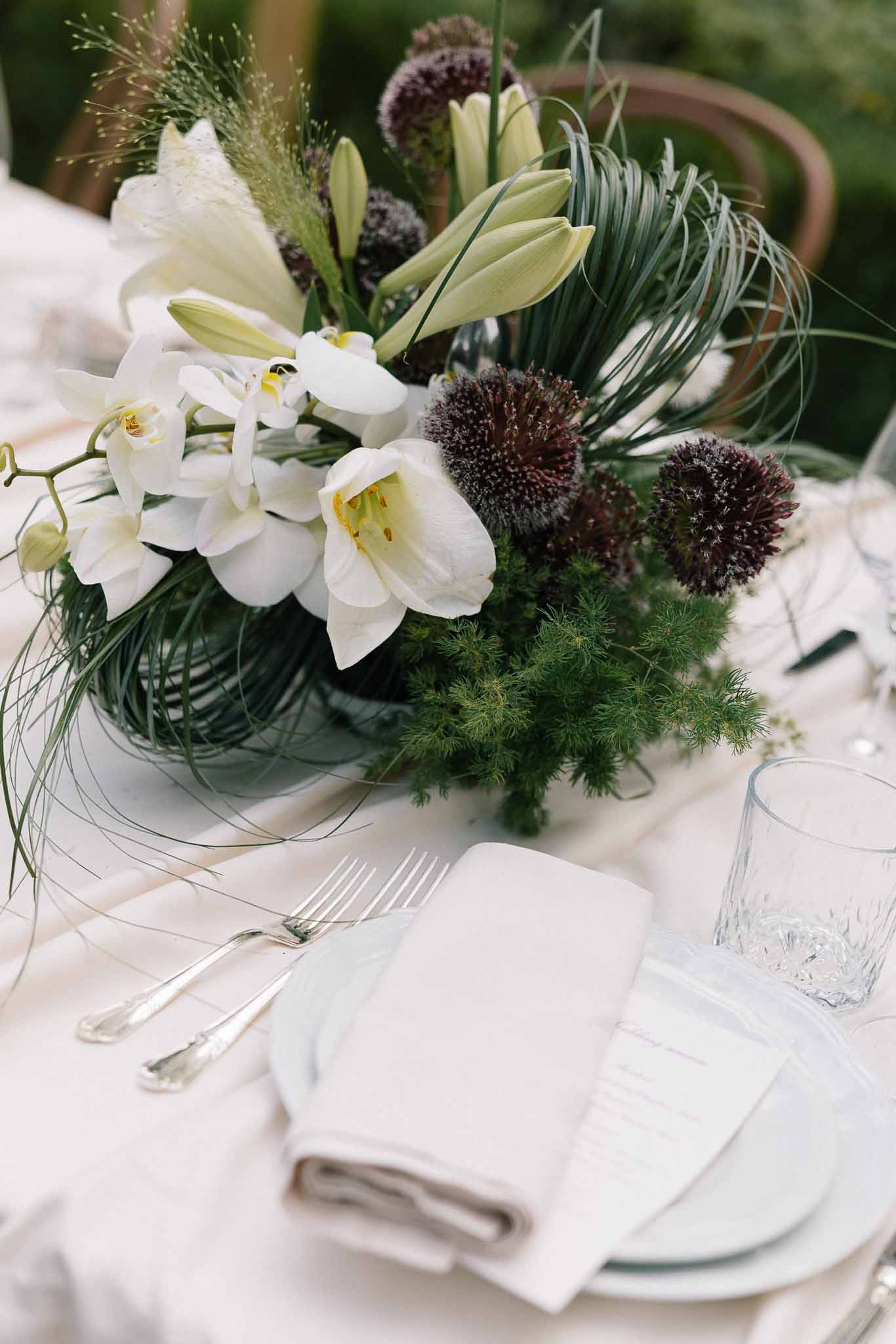 Reception centerpiece of white amaryllis, orchids, and purple allium in dark green bowl with silver place setting