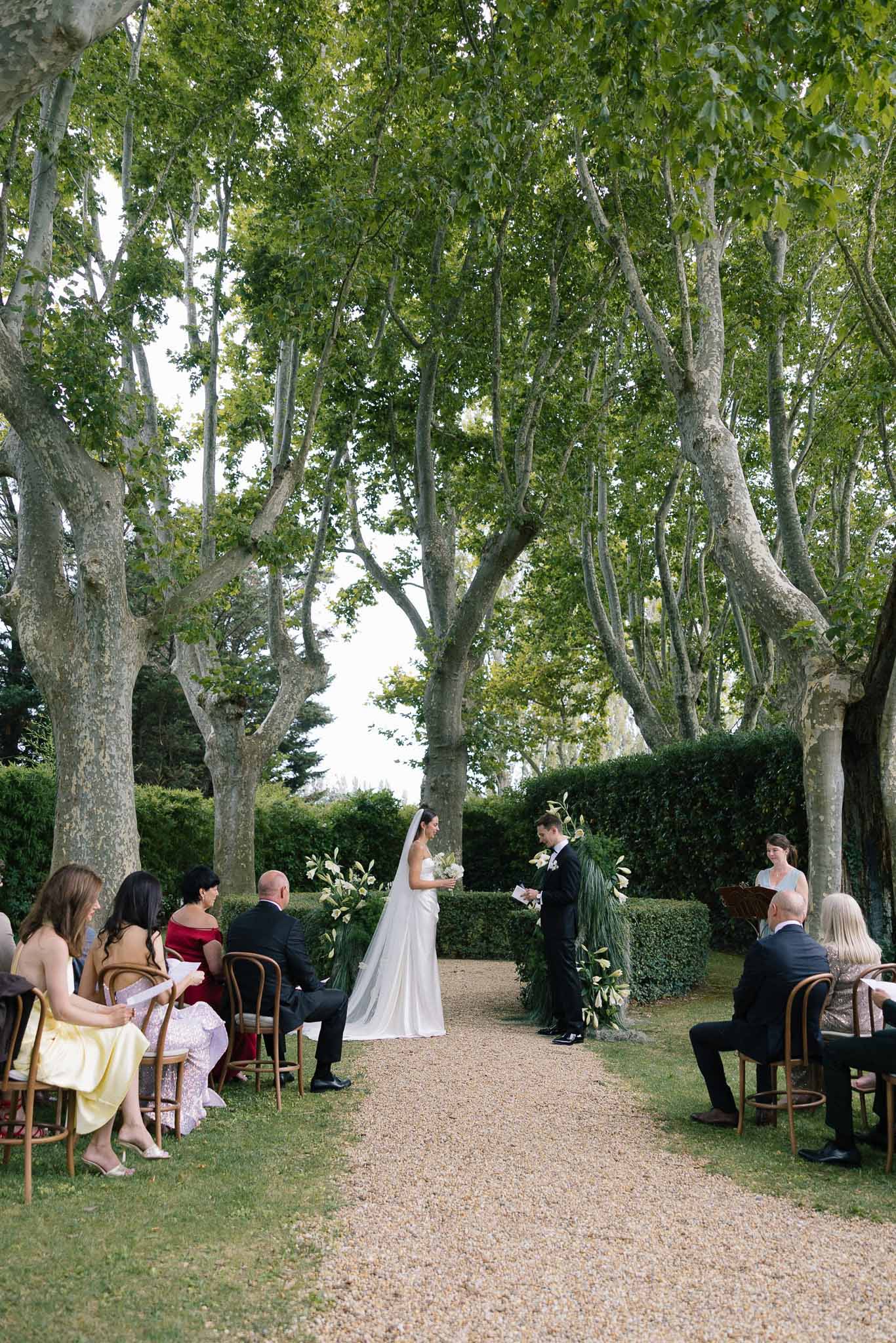 Vow exchange under plane trees with white and tropical green floral arrangements on gravel allee