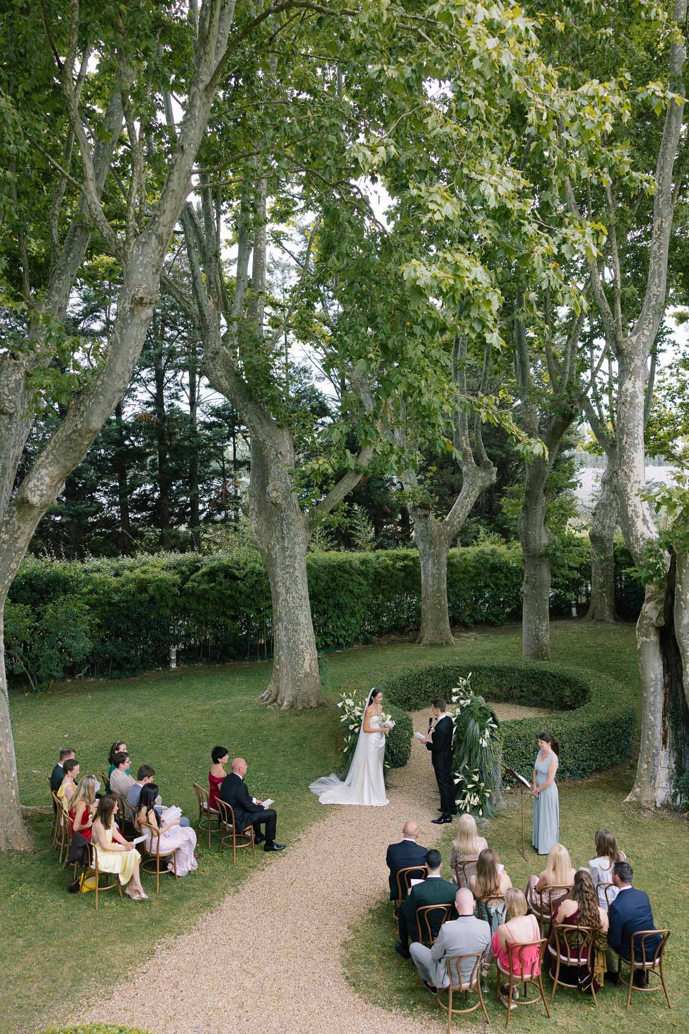 Outdoor garden wedding ceremony with couple at altar, white lily arrangements, and 25 seated guests on bentwood chairs