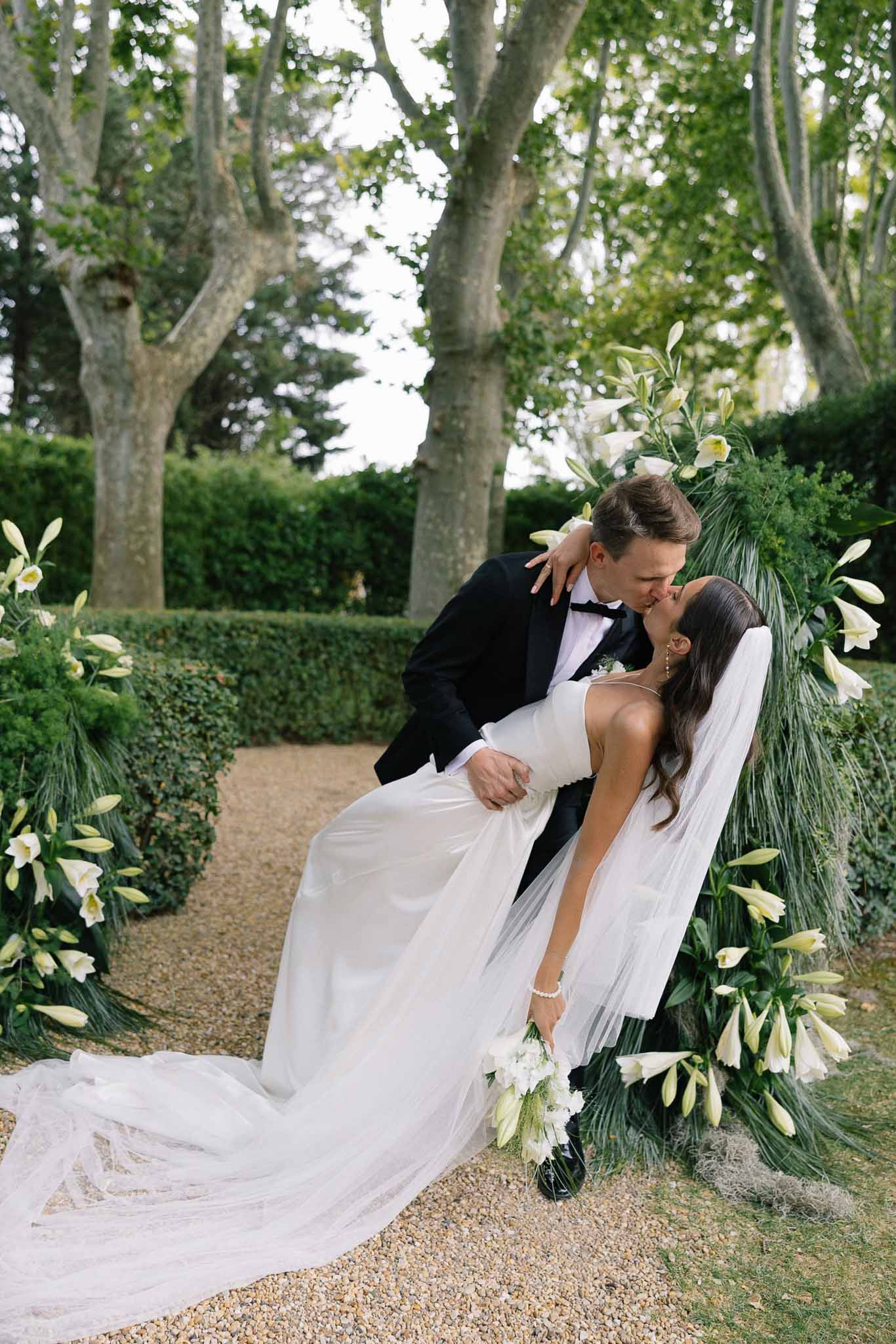 Bride and groom dip kiss before white lily arch with palm fronds and matching ground arrangements in garden