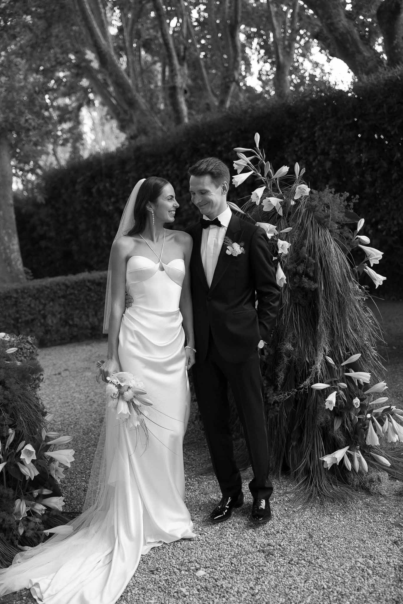 Black and white portrait of bride and groom on gravel path with tall sculptural floral installation behind them
