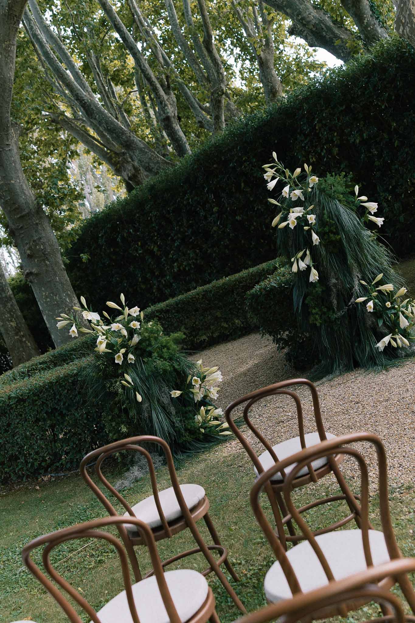 Circular floral arch of white lilies and dark foliage with bentwood chairs before clipped hedgerows