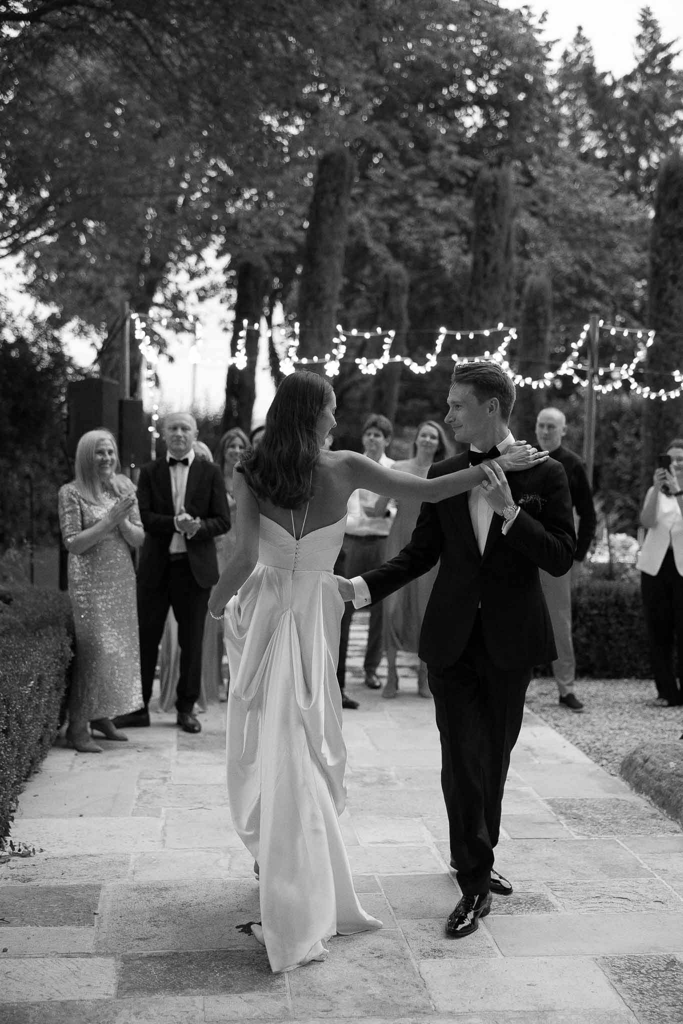 Black and white first dance on stone terrace with fairy lights ten guests watching and trimmed hedgerows