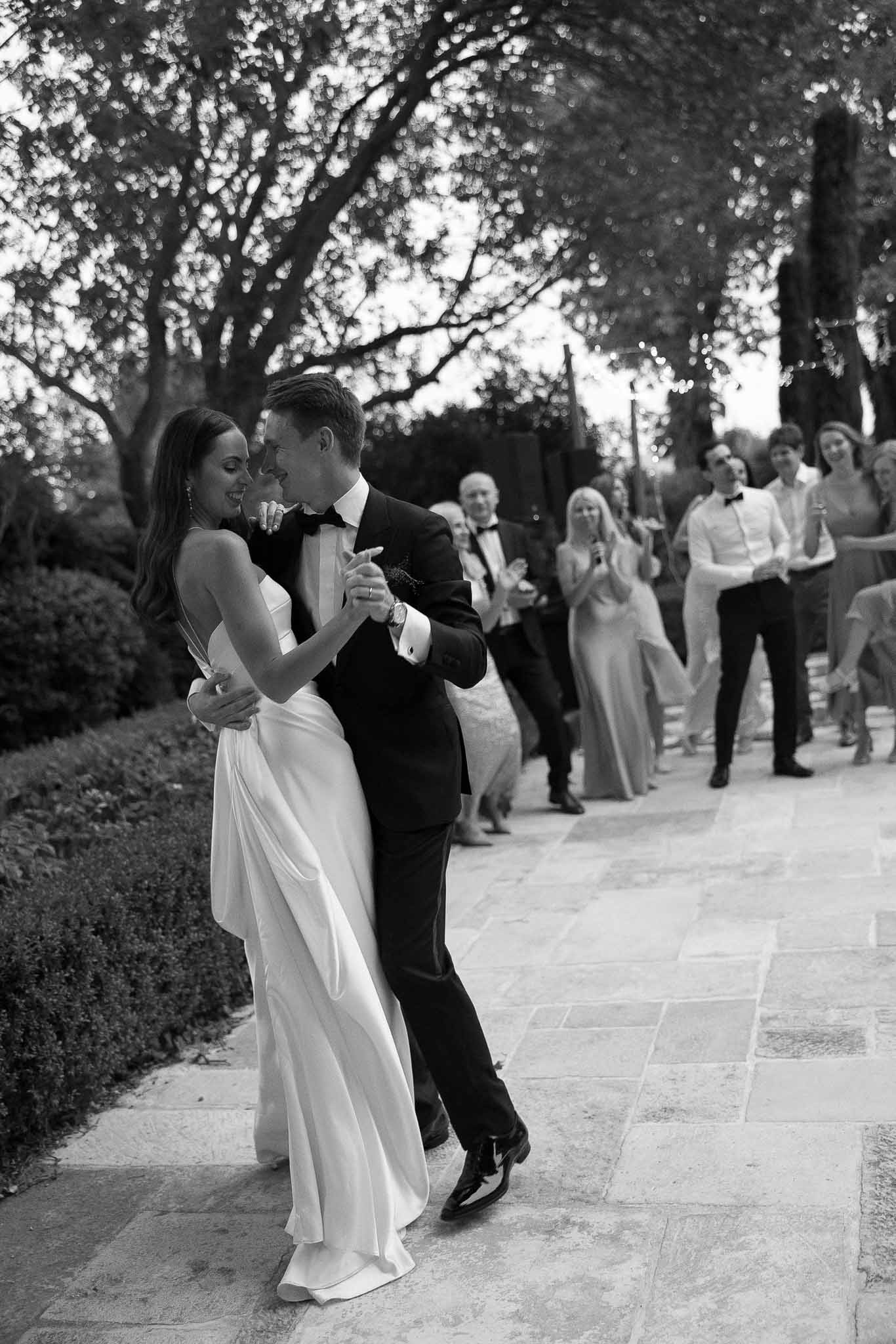 Bride in satin slip gown and groom in tuxedo share first dance on stone terrace with guests behind in B&W