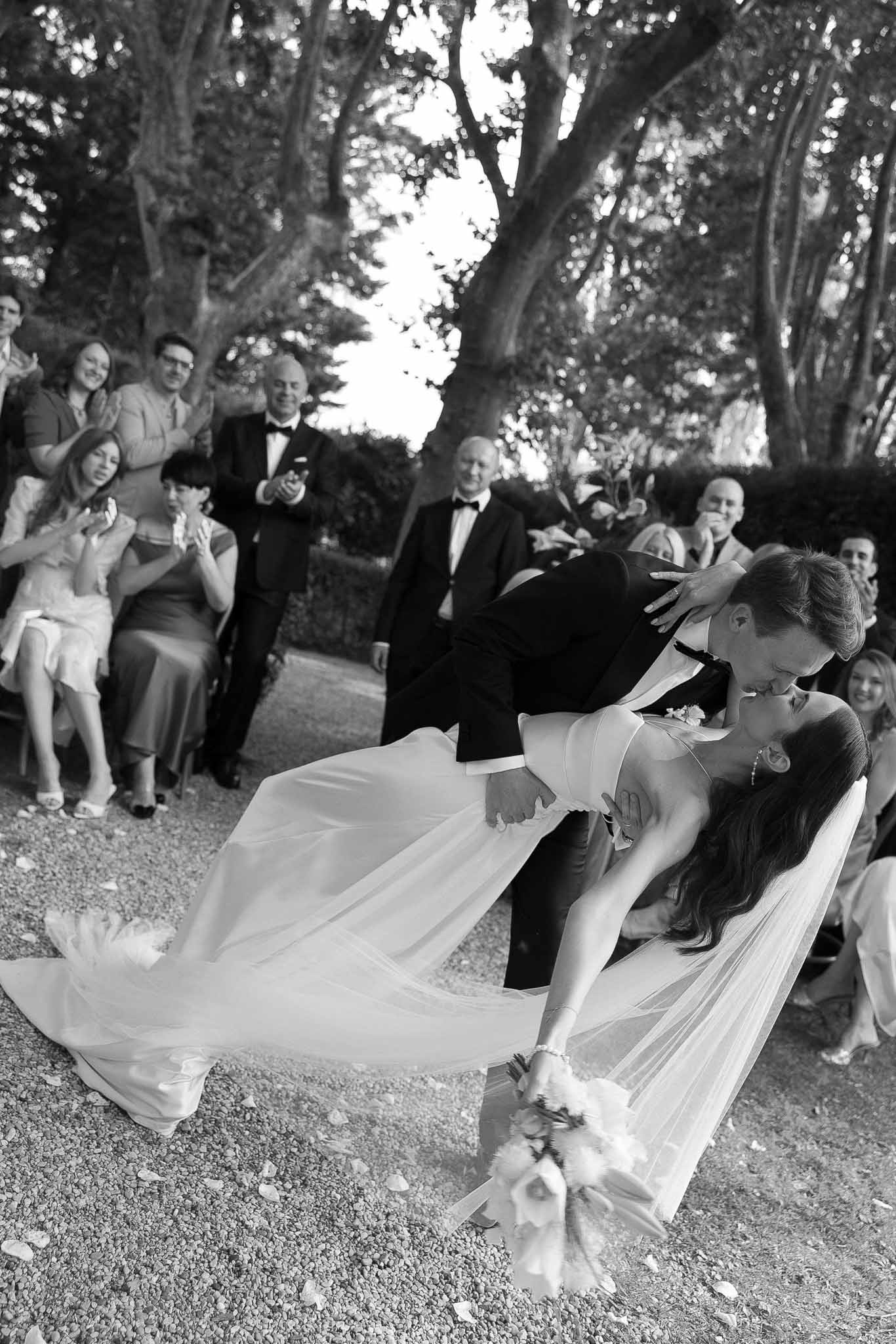 Black and white first kiss dip with veil fanning across gravel as guests applaud at garden ceremony