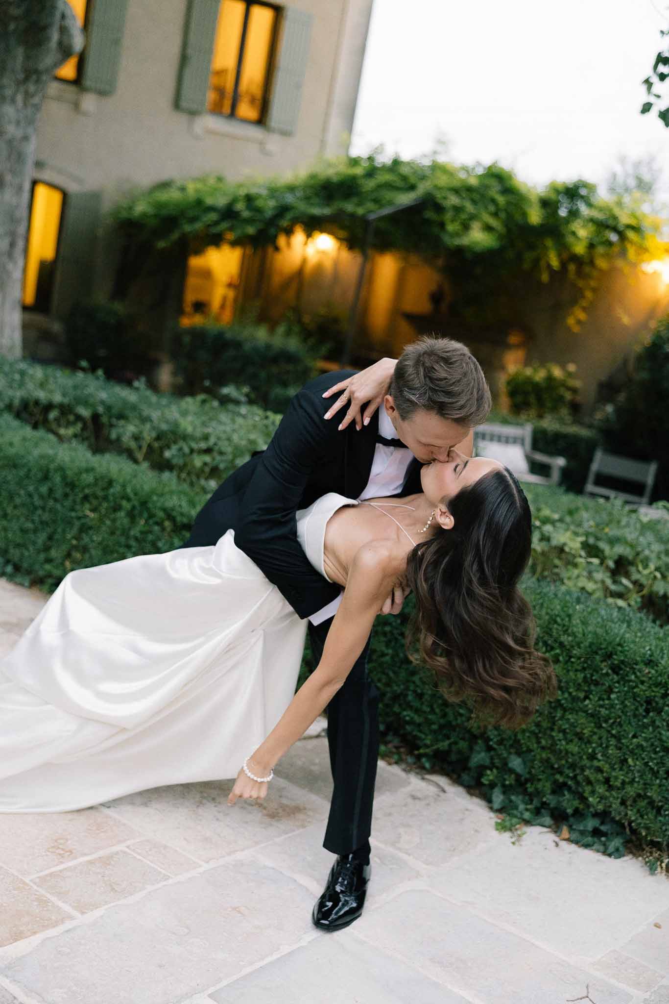 Groom dipping bride into kiss at dusk on stone terrace with manor house glowing behind