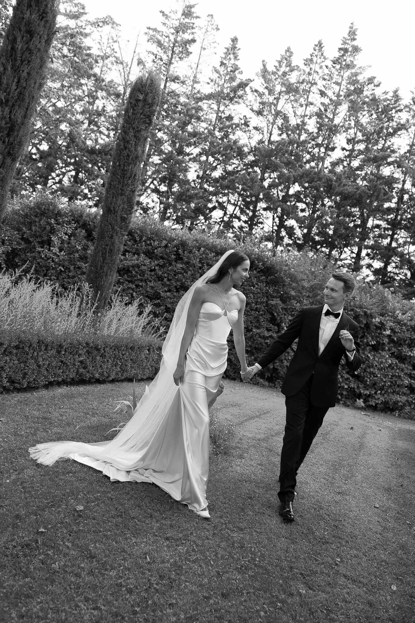 Black and white photo of bride and groom walking hand in hand in a garden