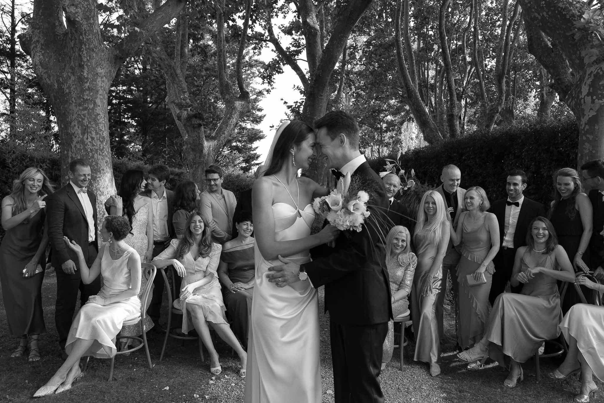 Black and white wide shot of bride and groom pressing foreheads together as guests clap after outdoor ceremony