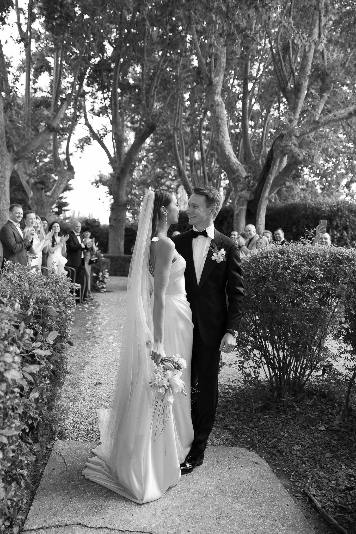 Black and white photo of couple kissing while walking back down hedge-lined aisle as guests applaud