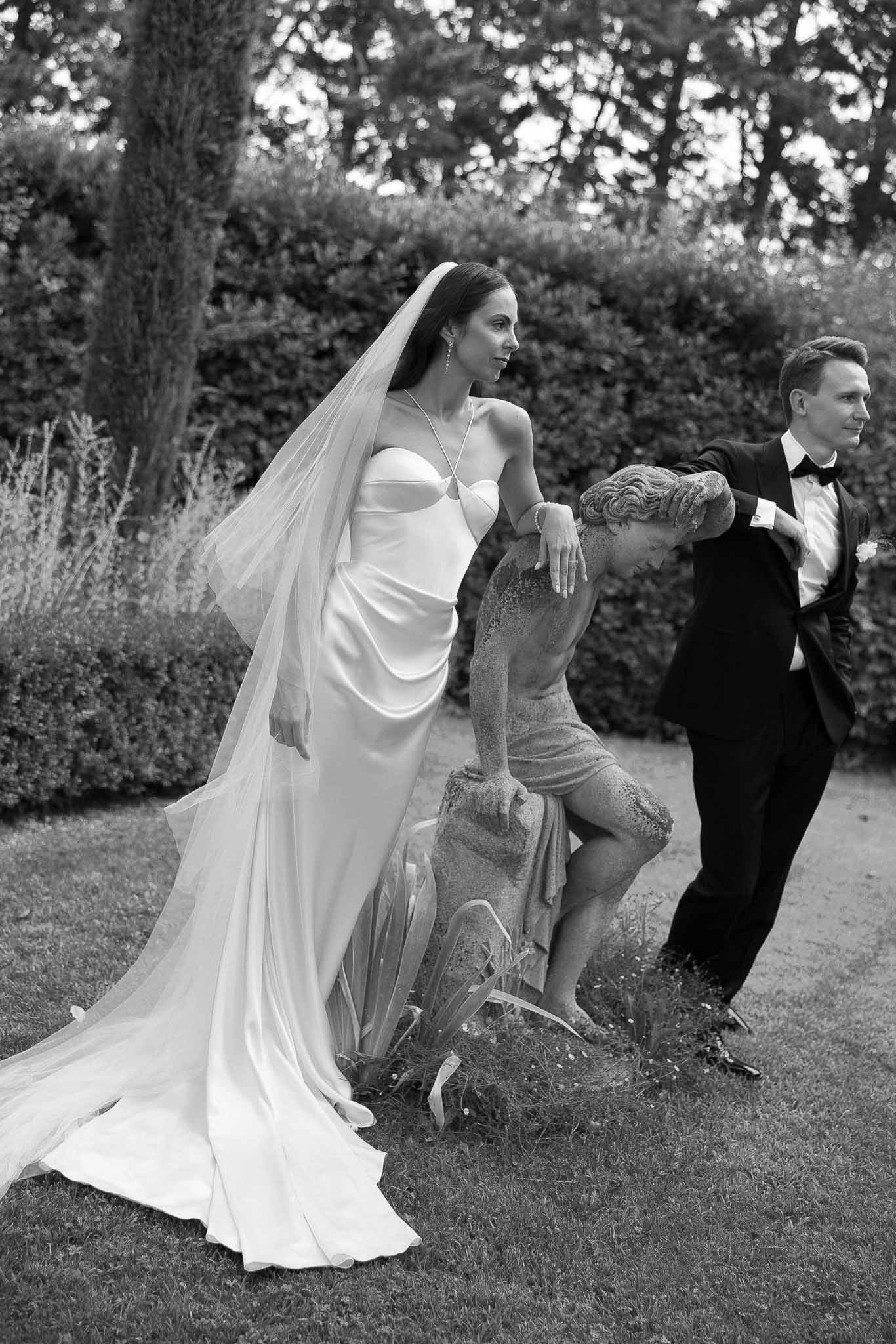 Black and white editorial of couple leaning on garden cherub statue, bride in satin slip gown and veil