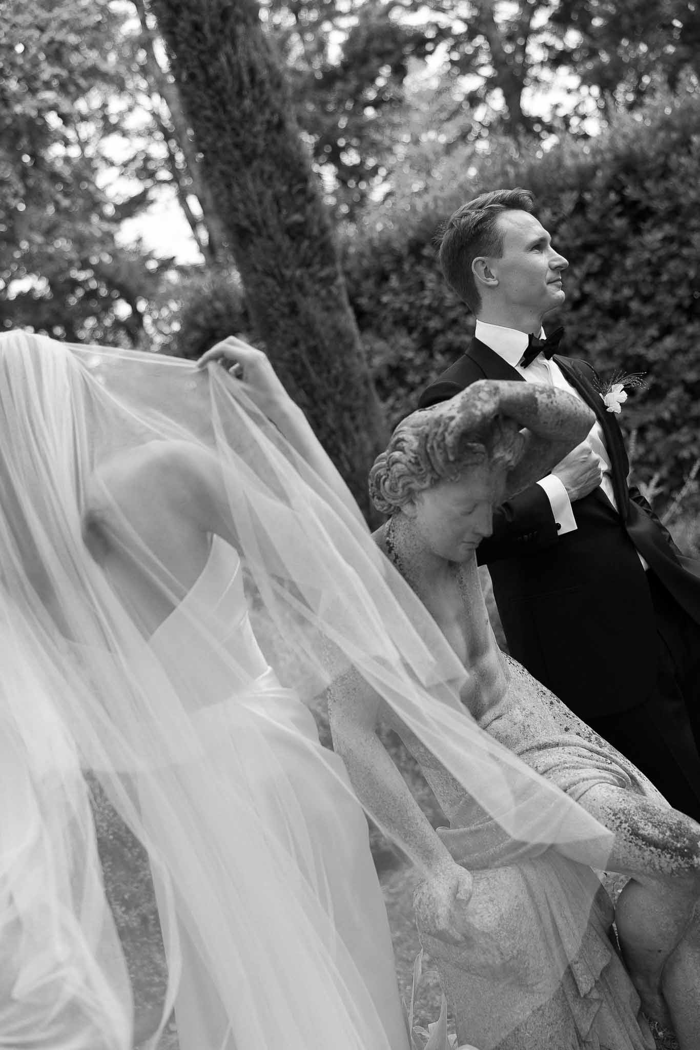 Bride billows cathedral veil beside groom and classical stone sculpture in formal garden in B&W