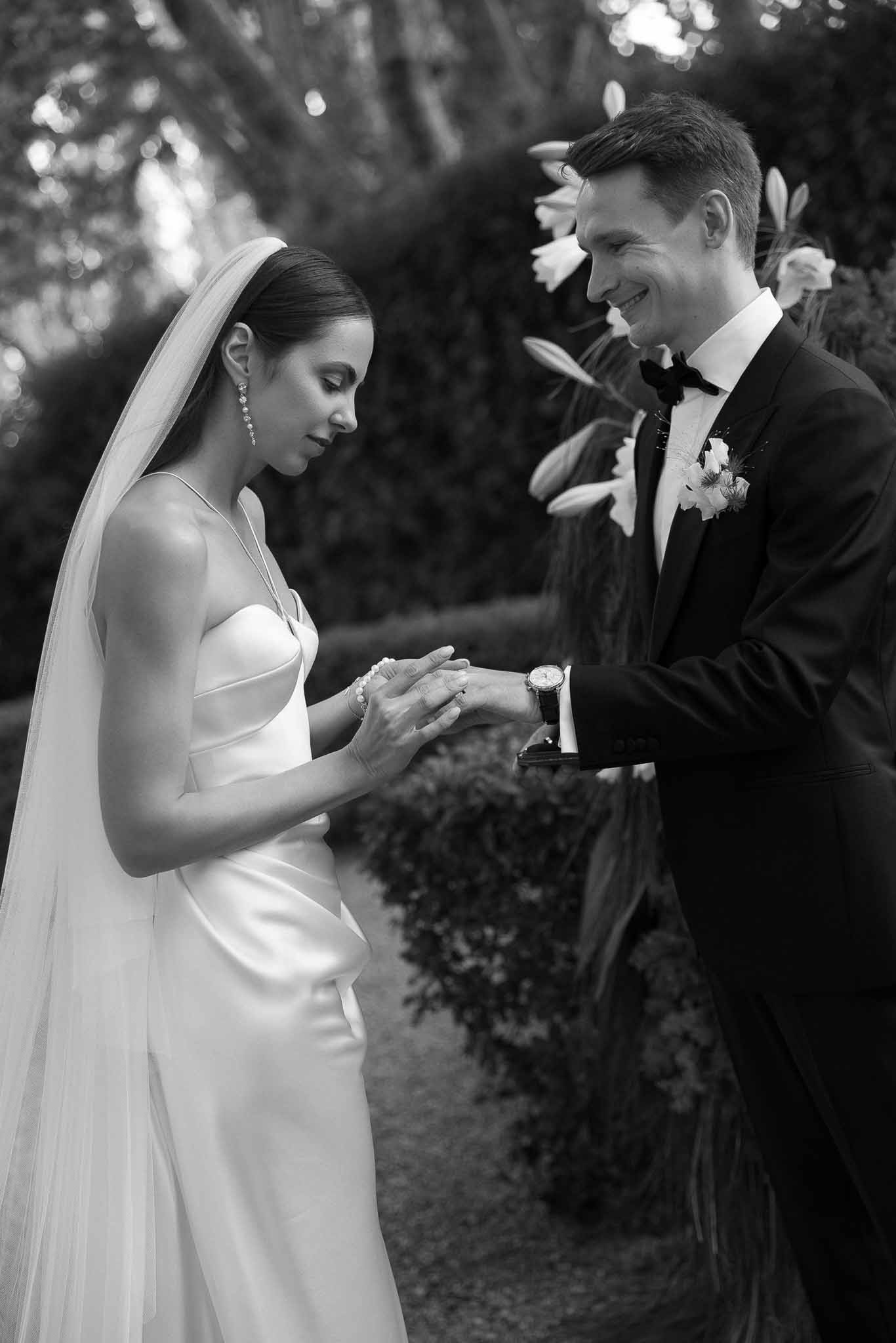Black and white portrait of groom placing ring on bride's hand during outdoor ceremony with lily backdrop