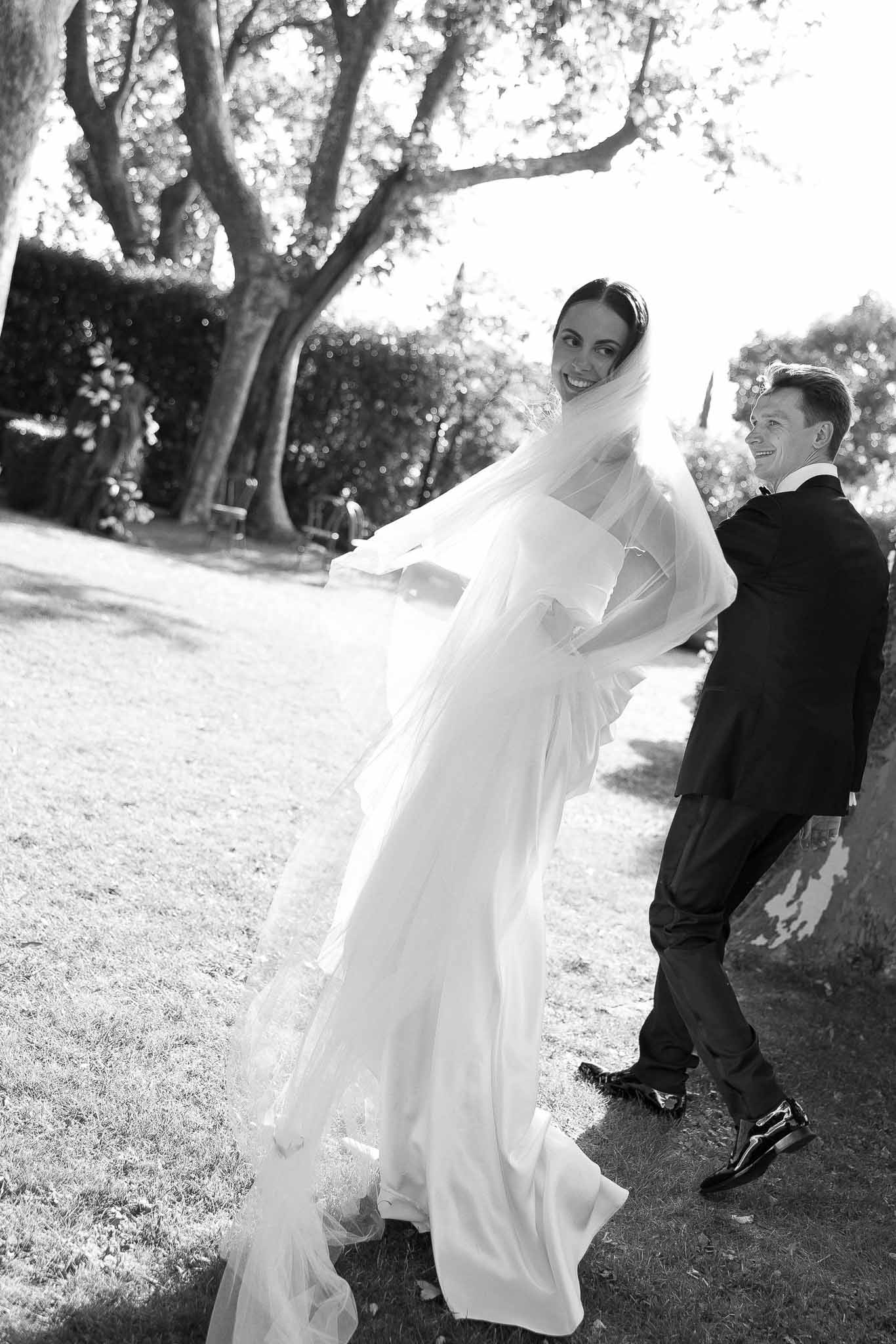 Bride with billowing cathedral veil and groom in suit walk through garden smiling in B&W