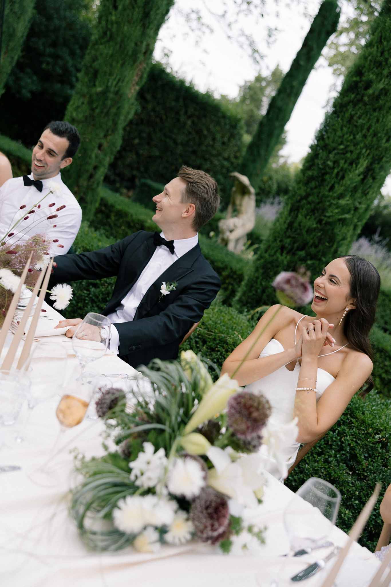 Bride and groom laughing at head table with burgundy allium and white gerbera centerpieces in formal garden