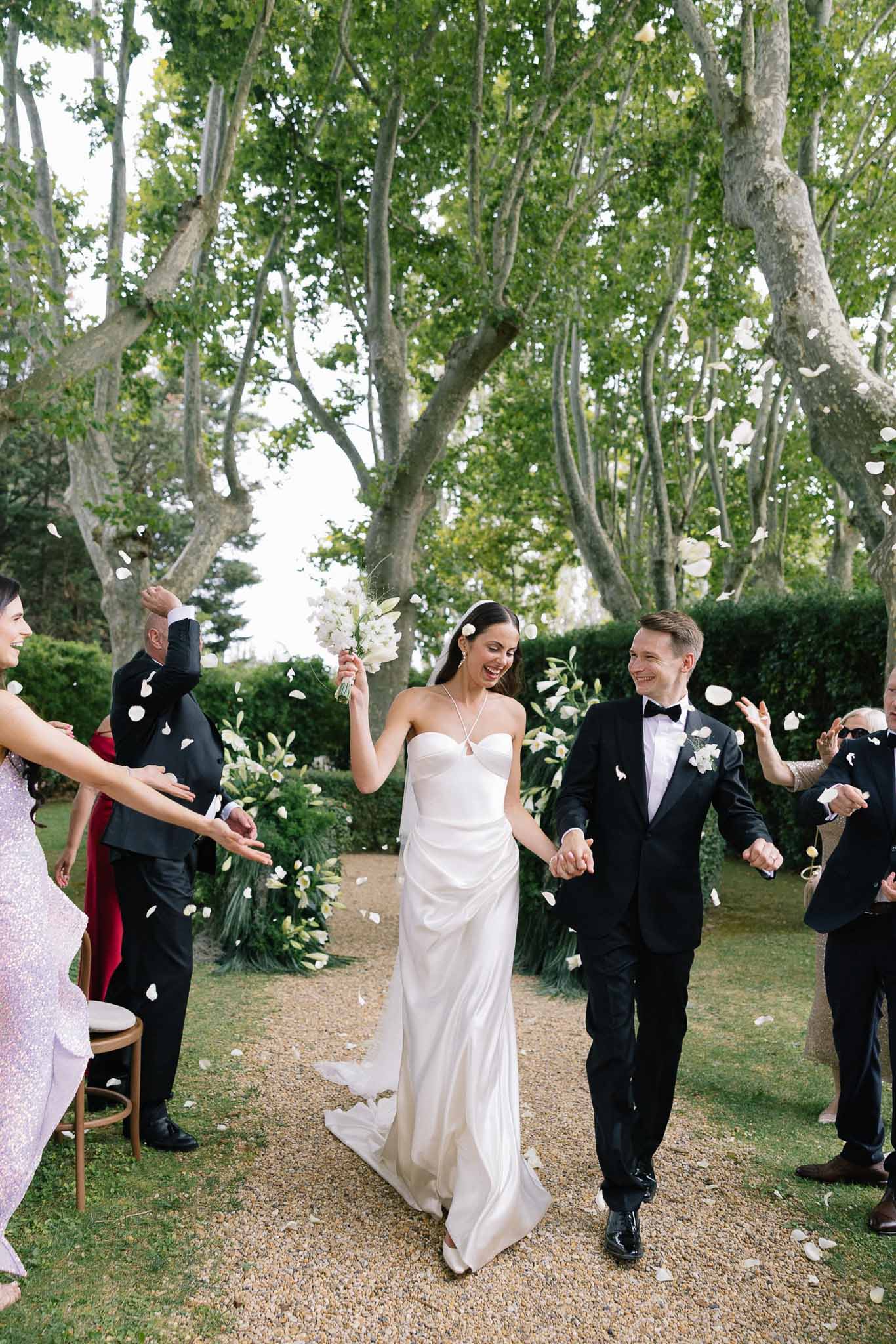 Bride in column gown with orchid bouquet and groom recessional through white petal toss under plane trees