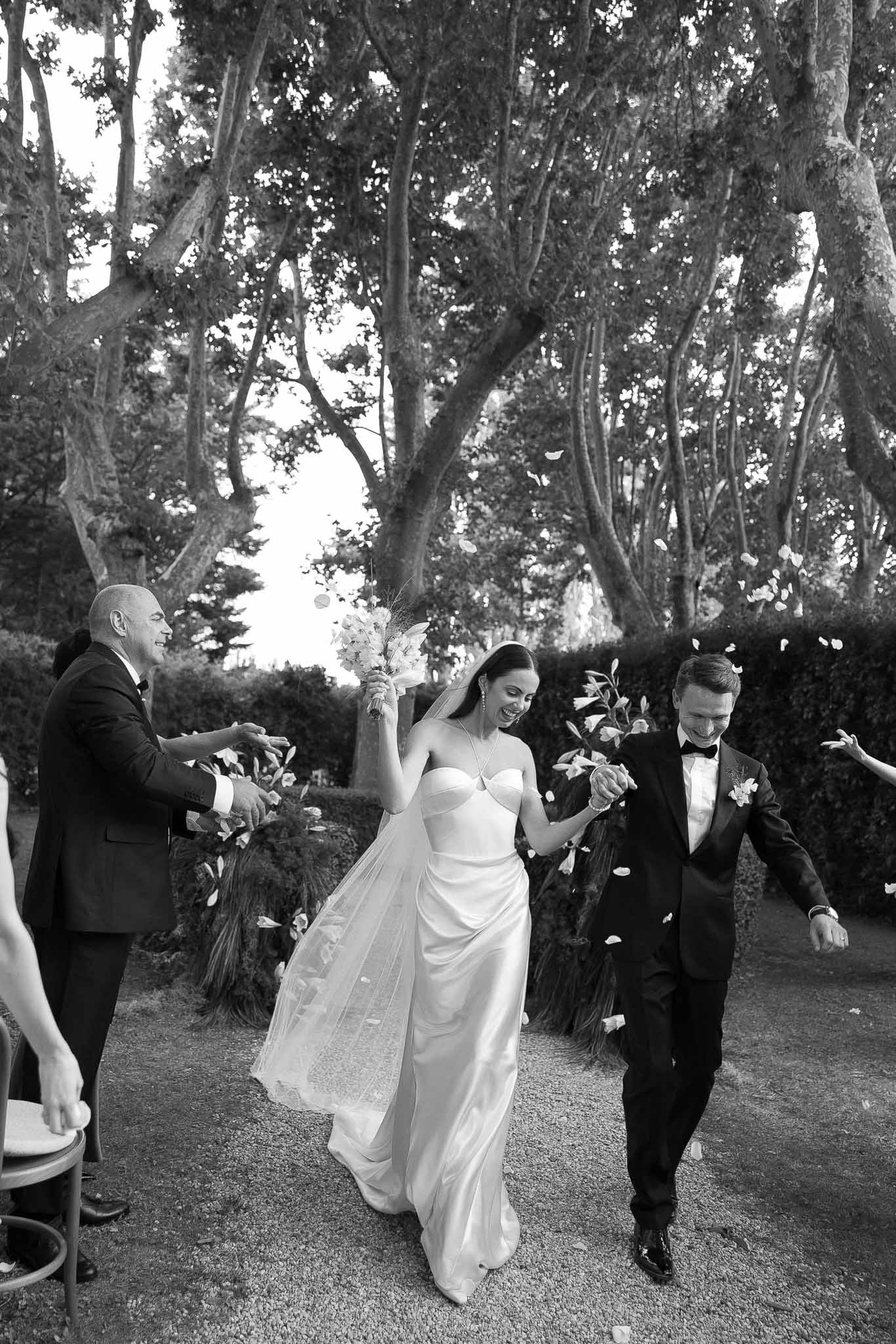 Black and white of couple laughing during petal toss recessional on tree-lined gravel path