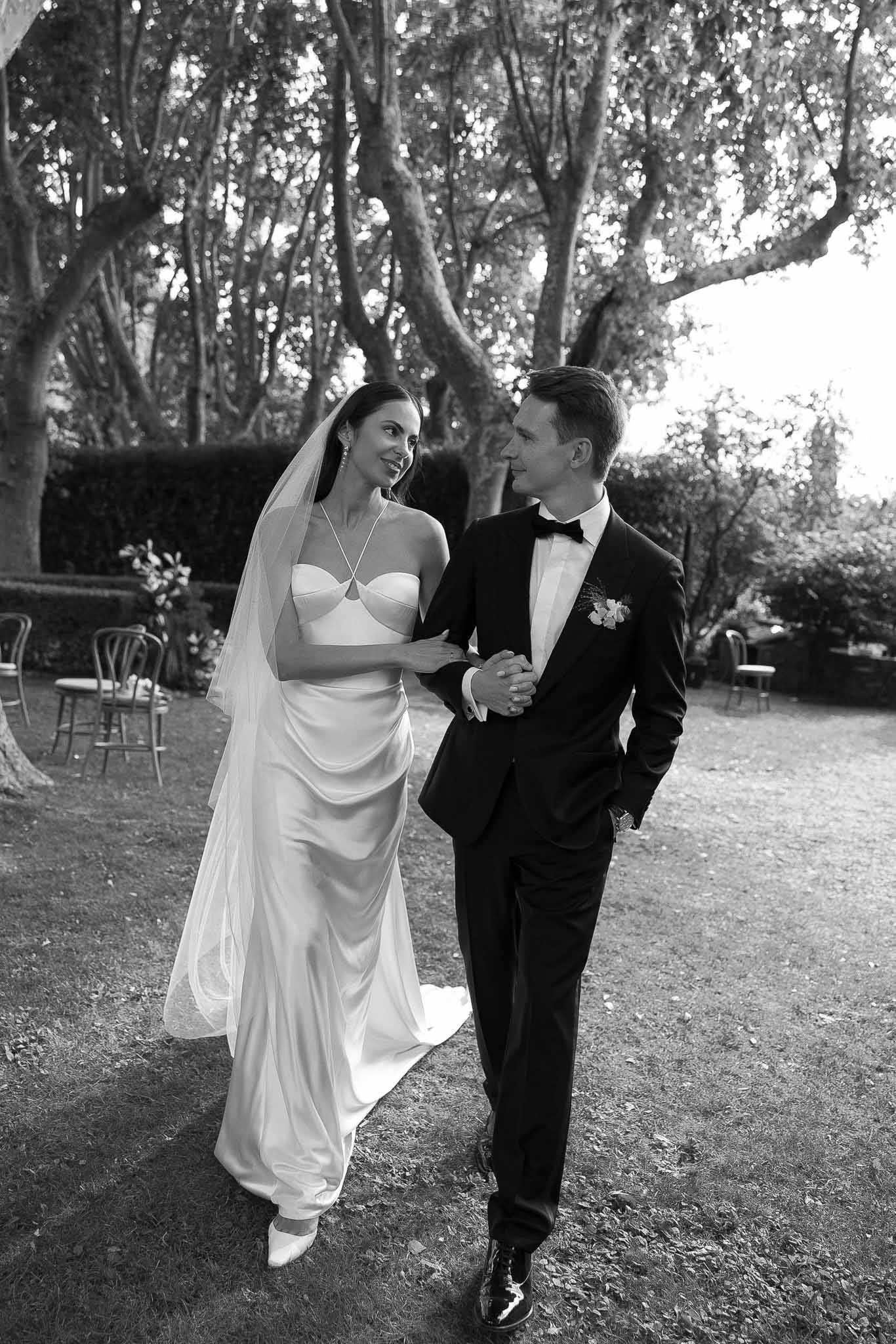 Black and white full-length portrait of bride in satin slip gown and veil walking with groom in tuxedo through garden