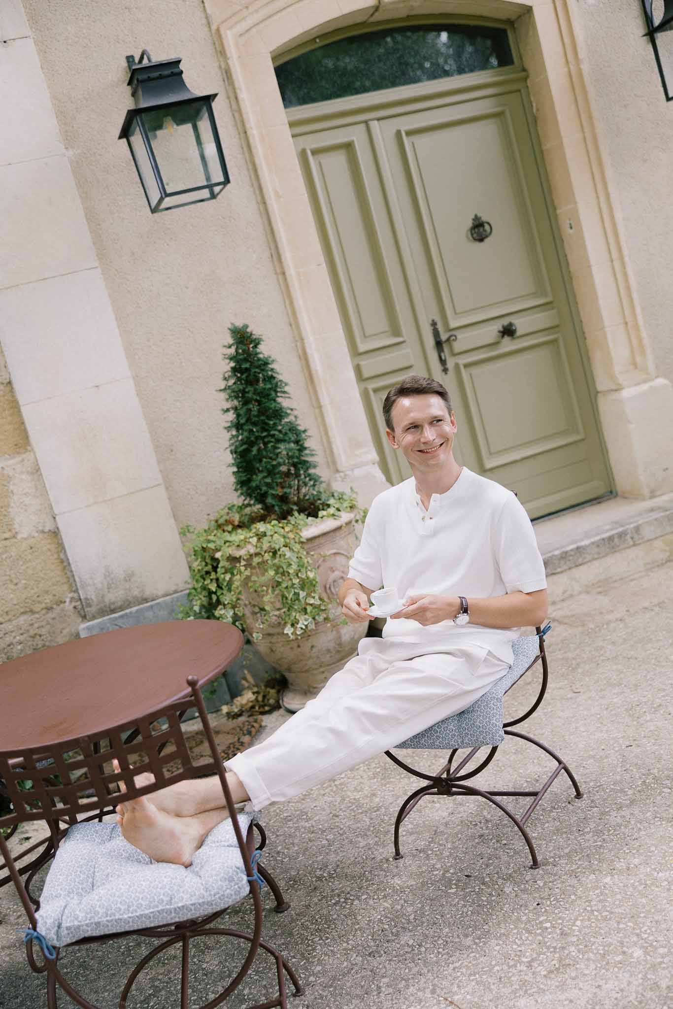 Groom in white linen shirt seated barefoot with espresso at chateau courtyard bistro table with sage green doors