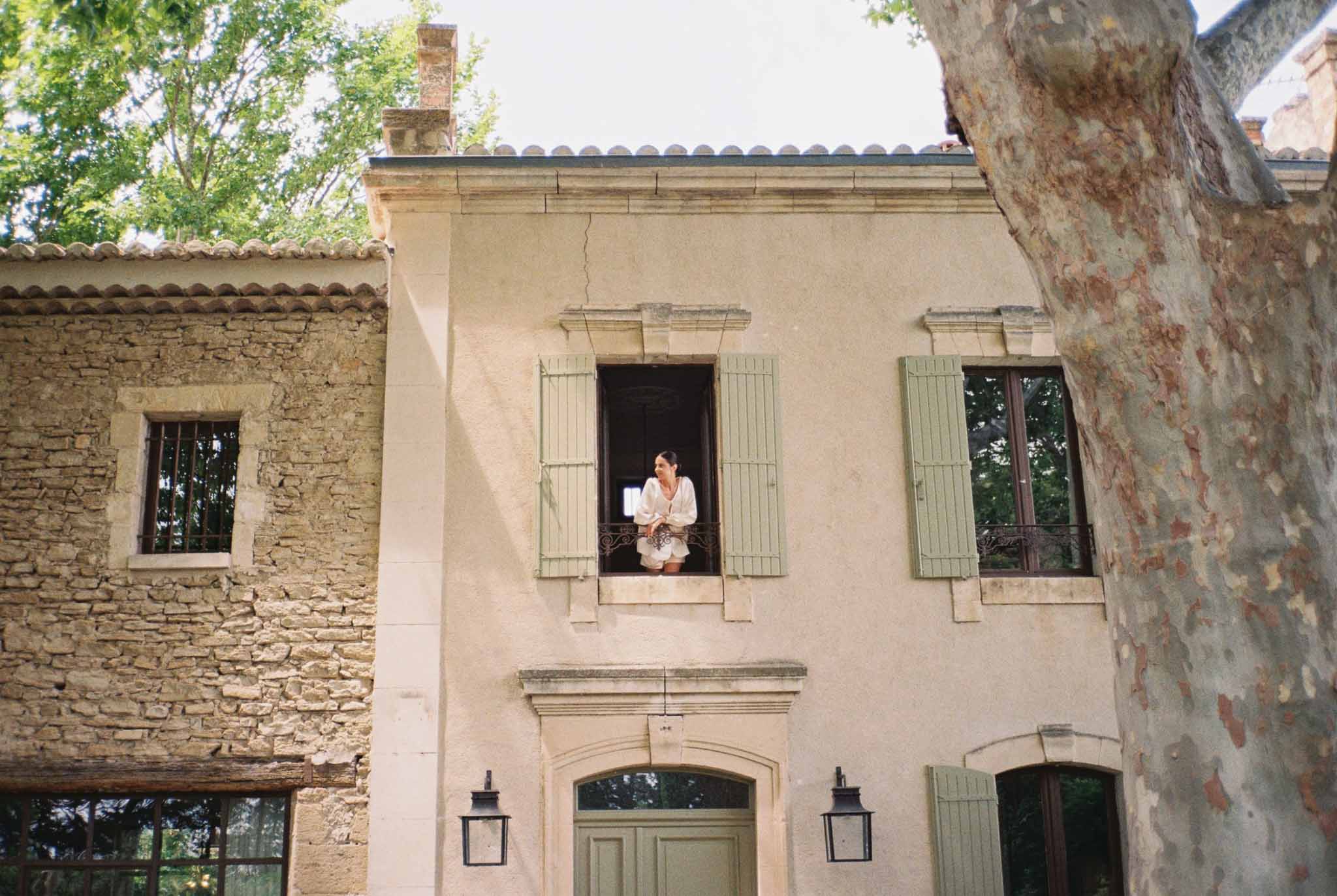 Bride in white robe leaning from second-floor window of Provencal mas with sage green shutters
